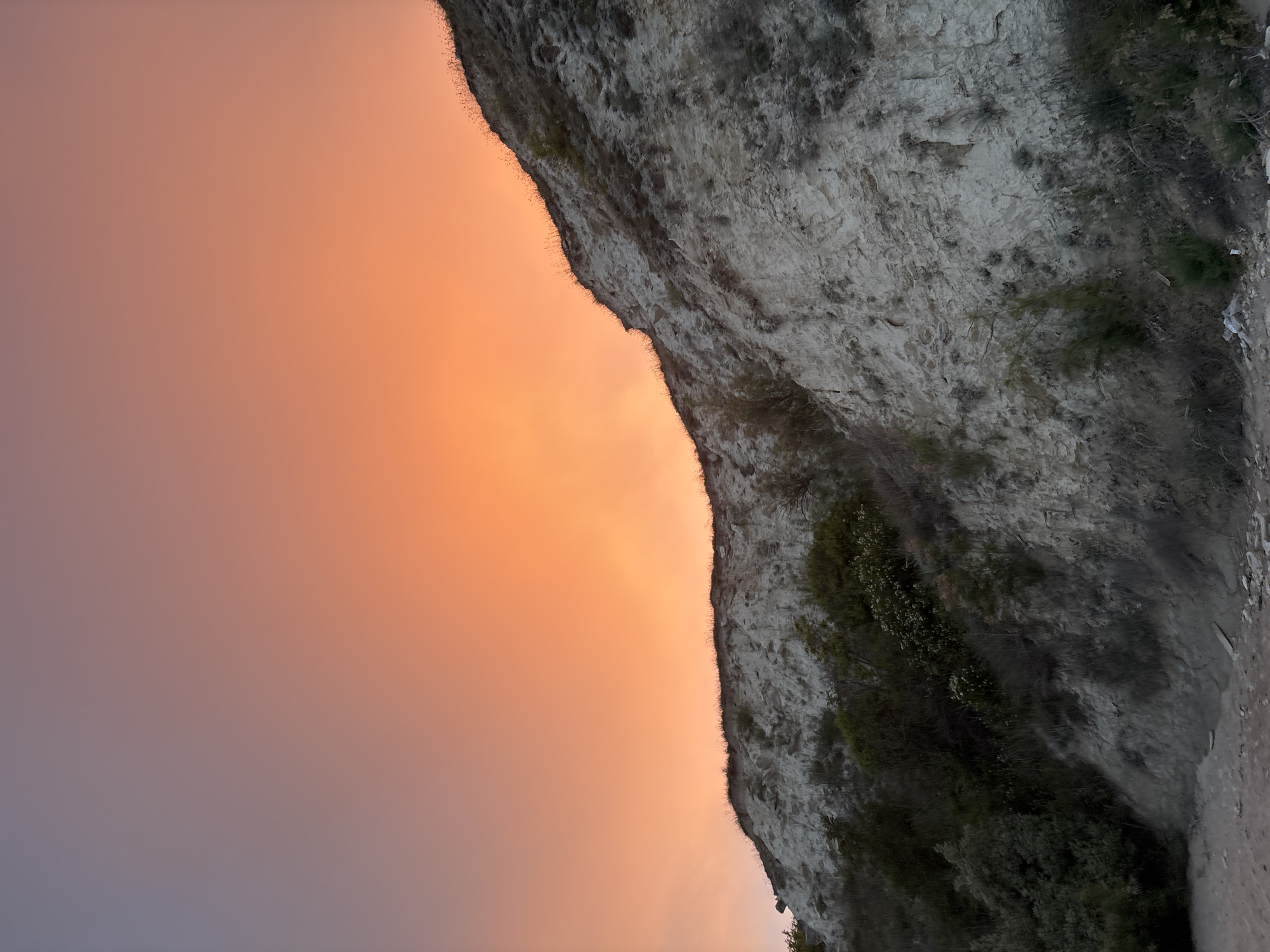 This is a landscape photograph taken in Topola, Bulgaria, featuring a rocky cliff face under a vibrant sunset sky. The sky transitions from a soft purple at the top left to a bright orange and peach hue towards the horizon, indicating either sunrise or sunset. The cliff is composed of light-colored rock with visible texture and striations, sparsely dotted with dry grasses and small, dark green shrubs. The lower part of the cliff is shrouded in shadow, and the very bottom of the image shows a sandy or gravelly ground with some scattered debris. No people are visible in the scene.