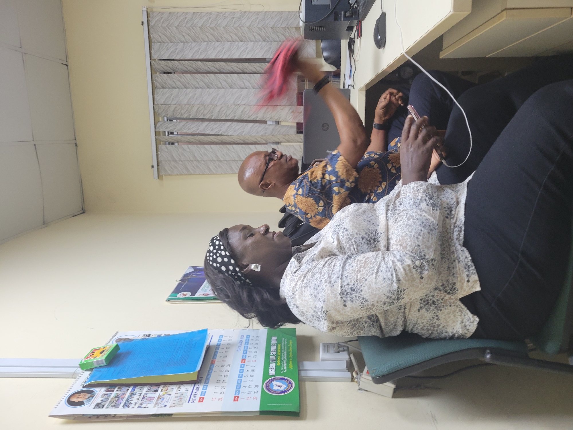 This image captures an indoor office scene, likely in Abuja, Nigeria, featuring two individuals engaged in various activities. In the foreground, a woman with dark skin, wearing a white polka-dotted headband and a light-colored patterned blouse with dark pants, is seated on a dark green office chair. She is engrossed in her light-colored smartphone, which is connected to a charging cable. Her gaze is directed downwards at the device.
Behind a light-colored desk, a man with dark skin, who is bald and wears glasses, is visible. He is dressed in a blue shirt with gold and brown patterns. His right arm is raised, exhibiting motion blur, as he holds a red item, possibly interacting with something above the desk, such as a computer monitor or equipment. A laptop (HP brand visible) and other computer peripherals like a keyboard and mouse are on the desk.
The office environment features light yellow walls and a window with light-grey patterned horizontal blinds in the background. Cables are visible around the desk area, indicating a typical workstation. On the wall near the woman, a calendar is displayed, showing dates for "NOVEMBER 2024" and "DECEMBER" (partially visible), along with the text "NIGERIA CIVIL SERVICE UNION (NATIONAL SECRETARIAT)". A blue notebook and a small green box are placed on the calendar. The overall scene depicts a functional office during what appears to be daytime, with both individuals focused on their respective tasks.