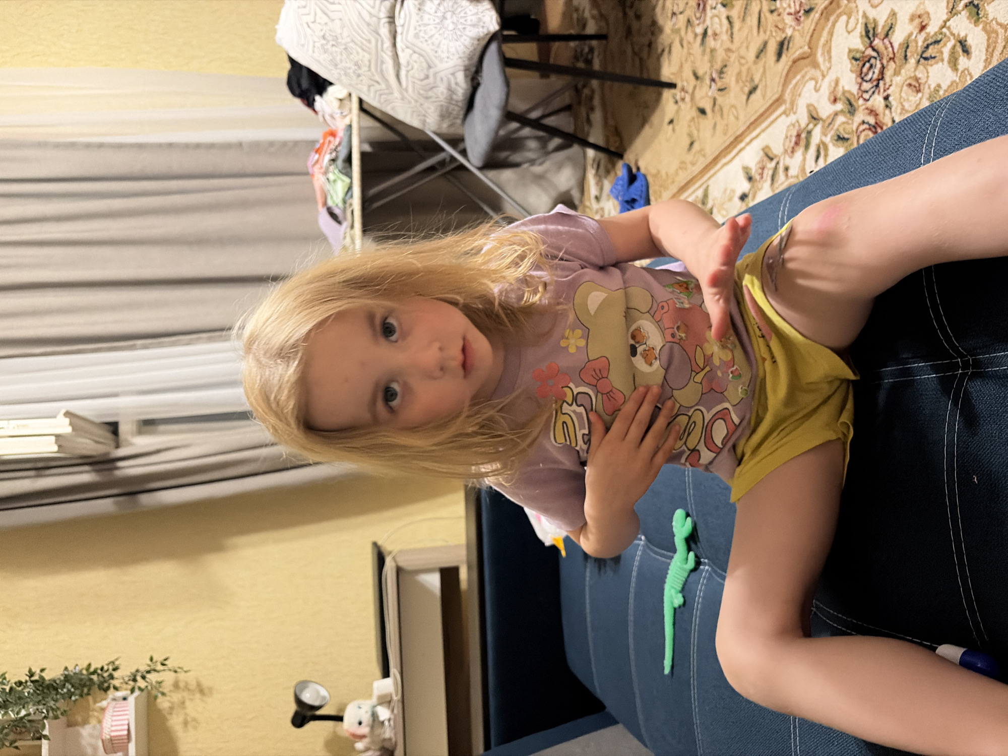 A young girl with blonde hair is lying on a blue couch. She is wearing a purple t-shirt with a cartoon bear graphic and yellow shorts. Her right leg is bent, and her hand is resting on her knee, where there is a small scrape. To her right, on the couch, is a green toy dinosaur. In the background, there is a rug with a floral pattern, a clothes rack with clothes on it, and what appears to be an ironing board. The lighting suggests it is indoors, possibly evening. The setting appears to be a home in Tambov, Russia.