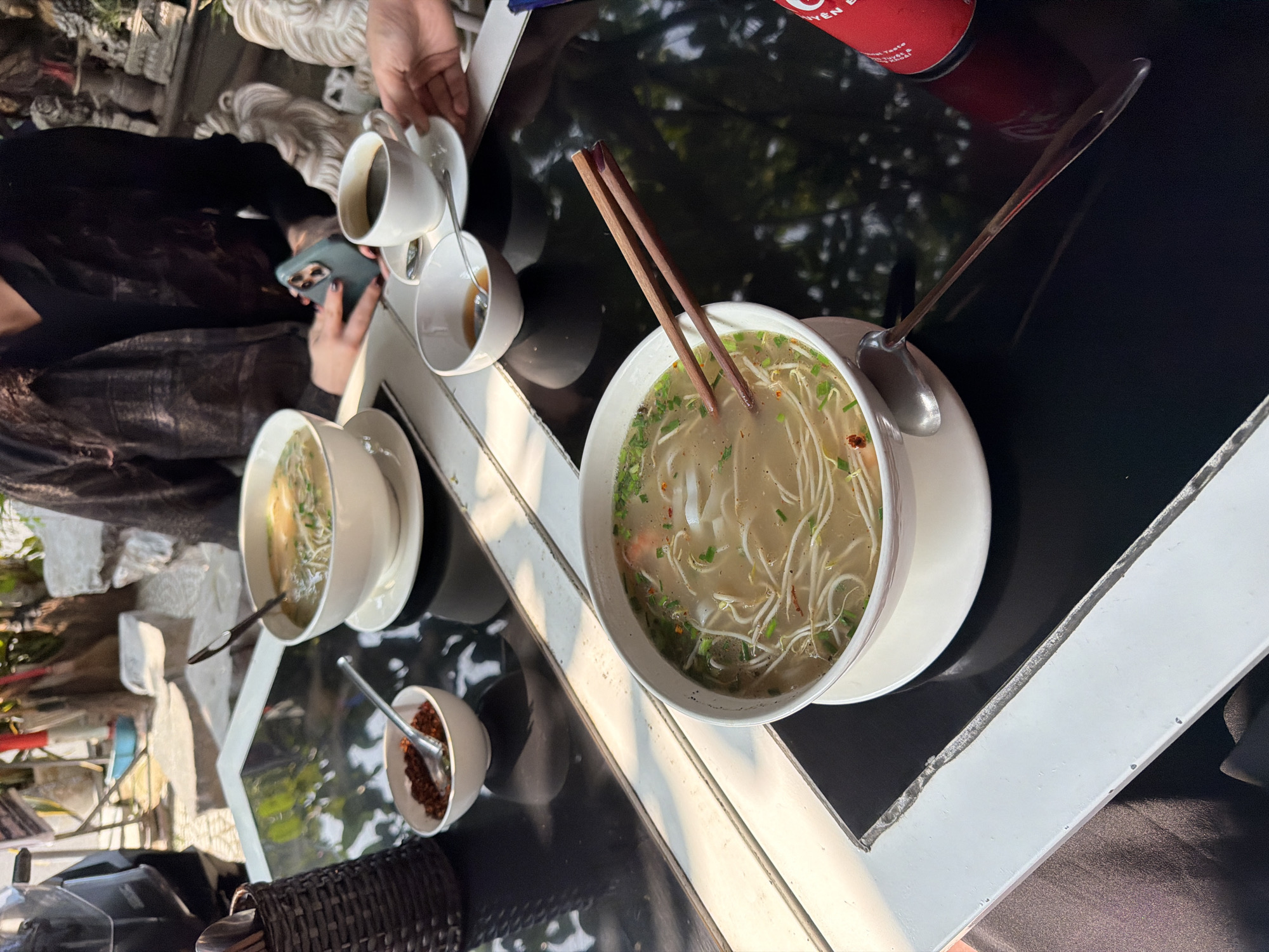A medium-angle shot depicts a restaurant table scene in Đà Nẵng, Vietnam. Two bowls of Vietnamese noodle soup (pho) are prominently displayed on a black table. A pair of chopsticks rests in one bowl, while a spoon is laid across the other. Condiments, including chili flakes, are placed near the soup bowls. Two cups of coffee sit towards the background, and a can is positioned to the right. A person can be seen using a smartphone. The setting seems to be an outdoor patio, with architectural elements and greenery visible in the background.