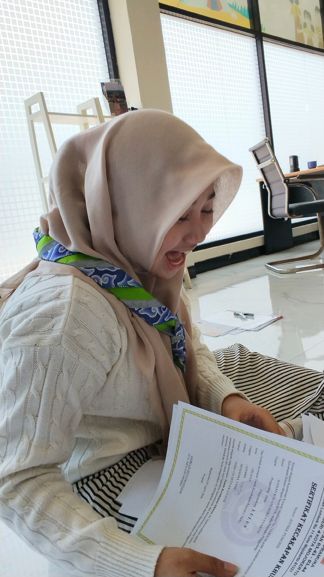 A young woman wearing a light pink hijab and a white cable-knit sweater is seated on a floor, looking down at a document. A patterned scarf with green, blue, and white designs is draped around her neck. She is holding the document with her hands and her mouth is open as if she is speaking or reading aloud. The document appears to be a certificate with Indonesian text, and some text is visible at the bottom right of the image. The background shows a large window with a grid pattern, a white chair, and a desk with office equipment. The floor is tiled and reflective. The text on the certificate includes "Mojokerto, 19 April 2016" and "NAMA: YUNI FATMA LESTARI". The certificate also contains the title "SERTIFIKAT KECAKAPAN KHUSUS" which translates to "Certificate of Special Competence". Based on the text on the certificate, the scene is located in Mojokerto, Indonesia.