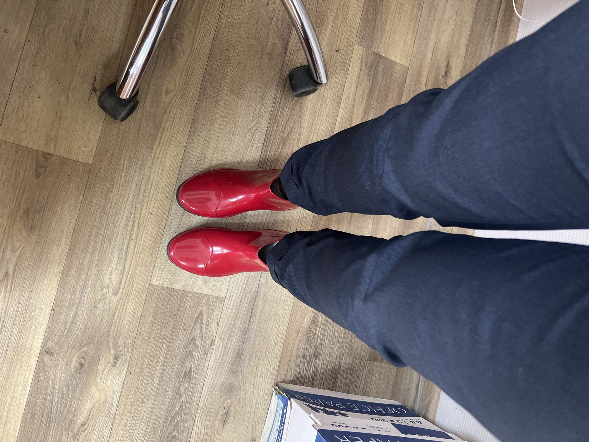 This is a first-person, overhead view of a person wearing dark blue pants and shiny red rain boots. The person is standing on a light brown wooden floor. To the left of the person is a cardboard box with "OFFICE PAPER" printed on it. To the right, the chrome leg and a black caster of an office chair are visible. The image does not contain information about the city of Volgograd, Russia.