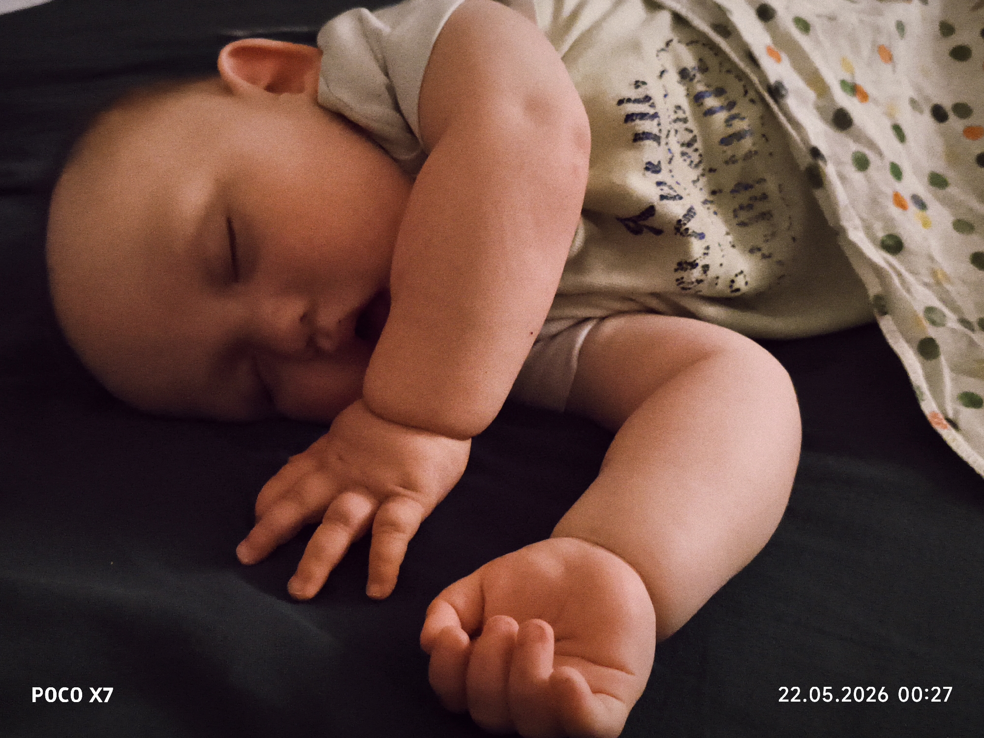 A baby is sleeping on a dark-colored surface. The baby is wearing a light-colored shirt and is partially covered by a blanket with a pattern of colored dots and a dark-colored design. The baby's face is turned to the side, and their arms are bent, with one hand open and the other slightly clenched. The image includes timestamps and camera information in the lower corners: "POCO X7" on the lower left and "22.05.2026 00:27" on the lower right. Location is unknown, Unknown.