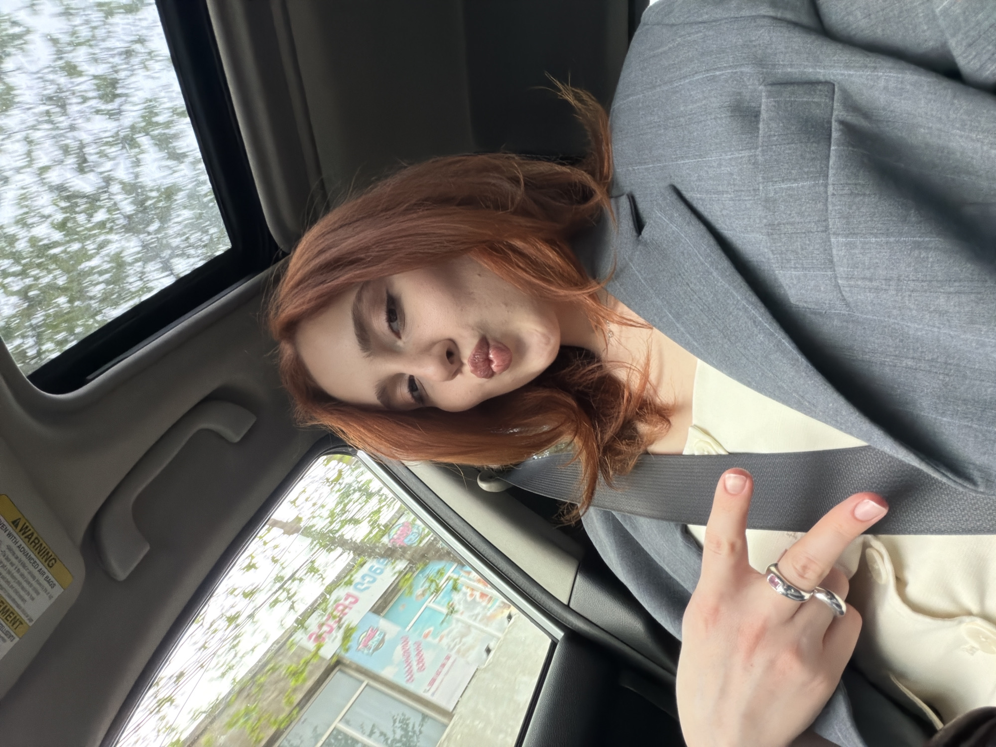 A young, auburn-haired woman sits in the passenger seat of a car, wearing a grey blazer over a light-colored shirt and making a kissing face at the camera. She is wearing a seatbelt and a large silver ring on her left hand, which is making a peace sign. Through the car window, a streetscape is visible, including what appears to be a sign for "Cherry Club 36" in Tbilisi, Georgia, suggesting the location. The car's interior is visible, including the roof and a warning label. The sky is visible through the car's sunroof.