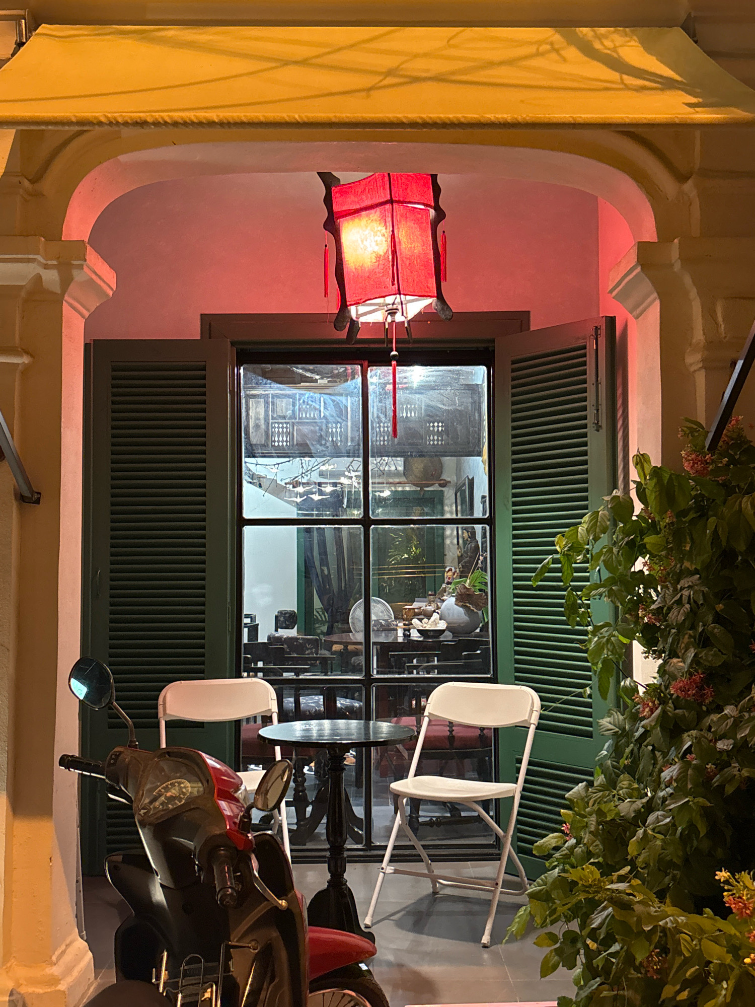 This is a nighttime, medium shot of an outdoor seating area in front of a closed establishment, possibly a cafe or restaurant, in Nha Trang, Vietnam. A red, square lantern with tassels hangs from an overhead yellow awning. To the left of the entrance, a dark red and black scooter is parked. Inside the establishment, visible through a large, multi-paned window, are tables, chairs, and a fan. Two white folding chairs are positioned outside the entrance, flanking a small, dark round table. Lush green foliage with small red flowers partially obscures the right side of the scene. The architecture suggests an older building with arched entryway.