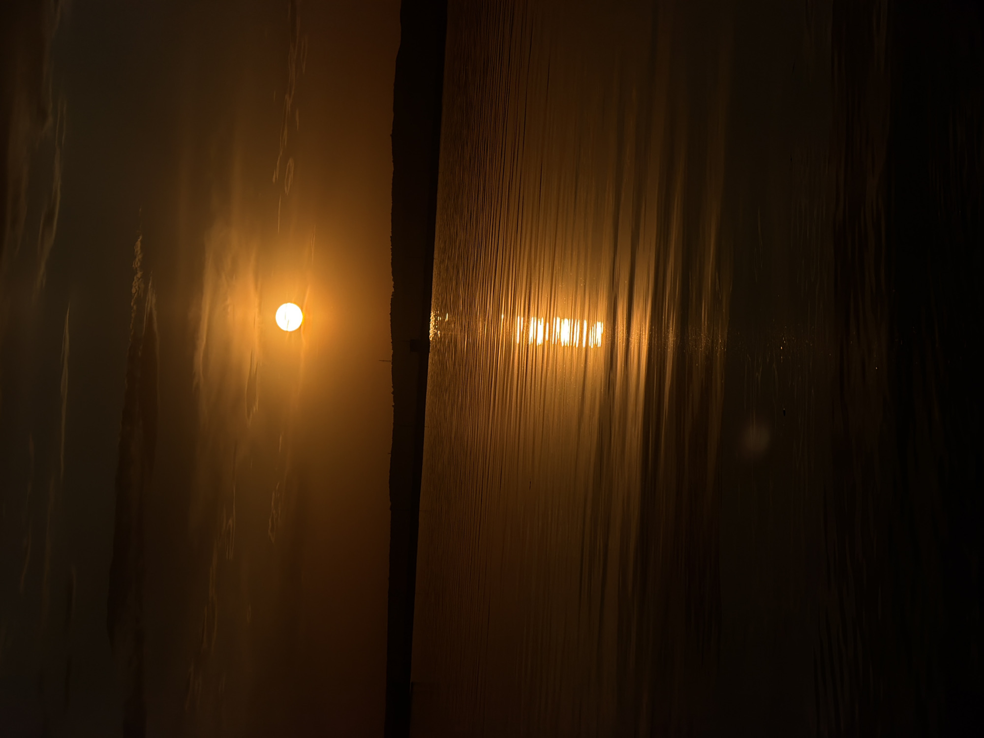 A low-angle photograph captures a scene illuminated by golden light. On the left, a bright, circular sun is visible through what appears to be a translucent or foggy environment. To the right, a dark, vertical strip divides the frame, leading to a textured, reflective surface that mirrors the sunlight in a series of horizontal bright lines. The backdrop is dark, with subtle vertical patterns suggestive of a curtain or other textured material. The location is an unknown city, state, and country. The overall composition is one of contrasts between light and shadow, and texture and smoothness.