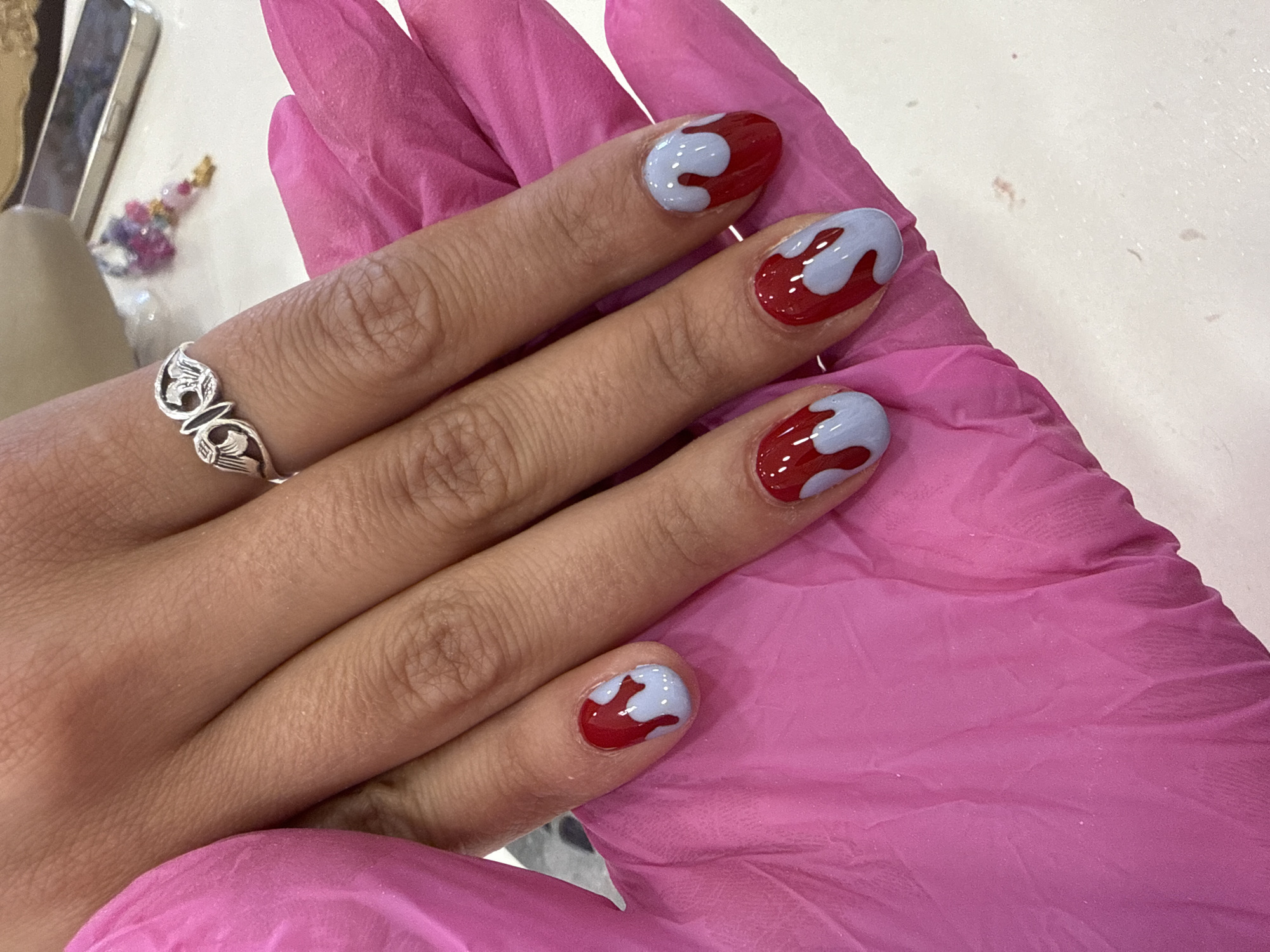 A human hand is resting on a pink latex glove-covered surface. The hand's fingernails are painted in a two-tone design featuring a red base with light purple or blueish-grey dripping accents. A silver ring with an abstract, possibly floral or avian design, is visible on the ring finger of the hand. The background consists of a white or light-colored surface. Some indistinct objects are visible in the upper left corner. There is no information to determine the location as Sofia, Bulgaria.