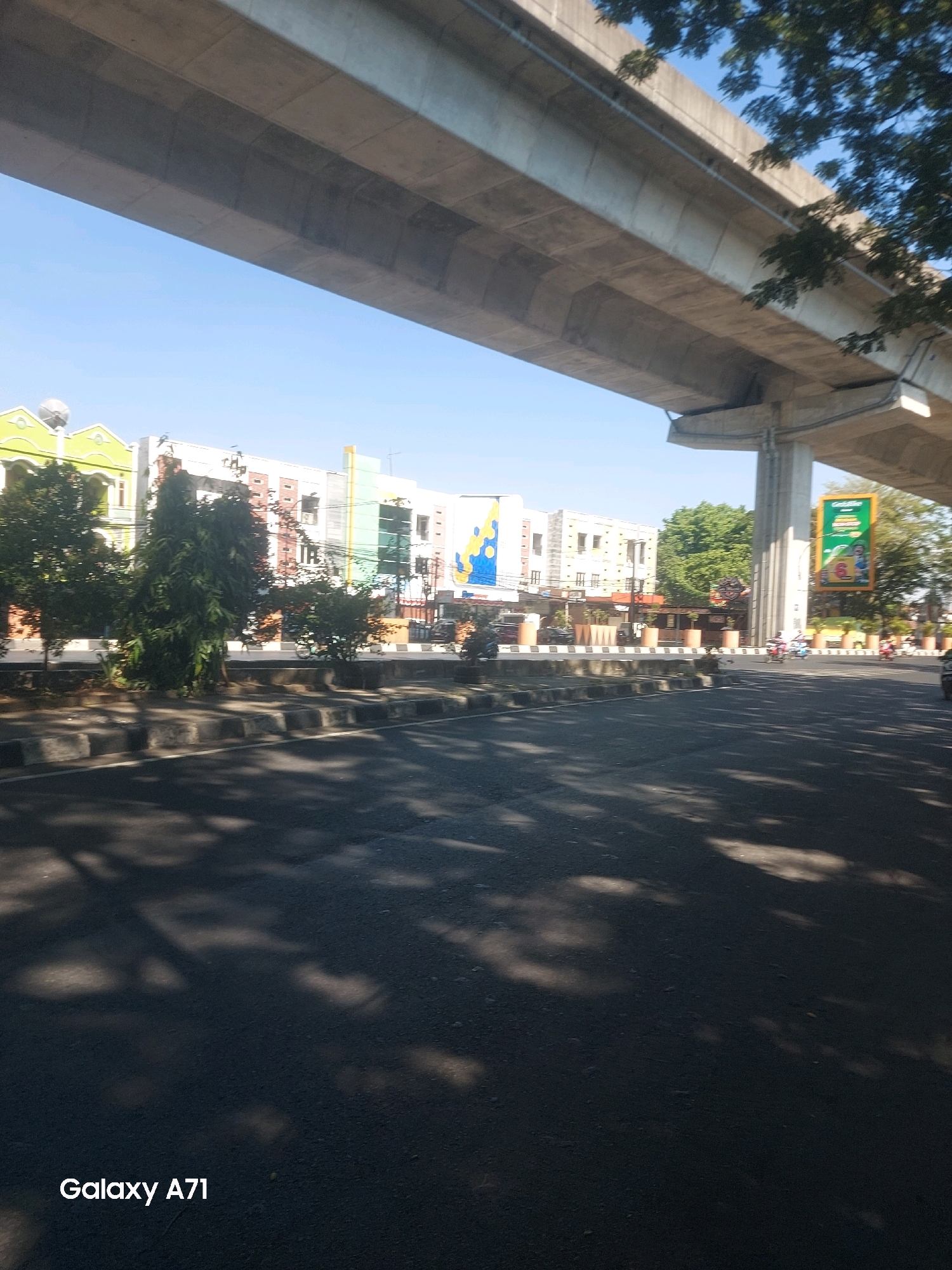 The image shows a street scene in Makassar, Indonesia, under an elevated highway structure. The highway is a prominent concrete structure spanning across the top half of the image. Below the highway, there are several buildings with colorful facades, possibly shops or offices. Trees line the sidewalk in front of the buildings, casting dappled shadows on the asphalt road. A few motorcycles and a yellow vehicle are visible on the road, suggesting traffic. A large green billboard advertising a burger with the number "6" is also visible on the right side of the image. The sky is clear and blue. The bottom left corner of the image contains a watermark "Galaxy A71".