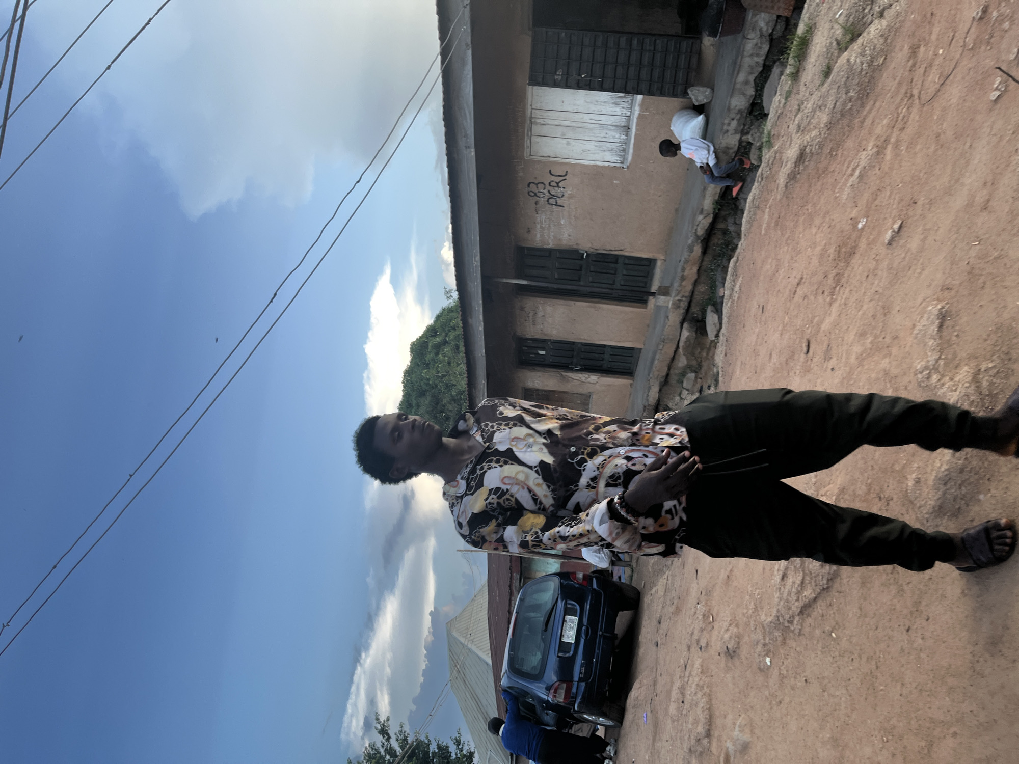A young Black male stands in the foreground, looking down and to his right. He is wearing a short-sleeved, patterned button-up shirt with a colorful design featuring swirling shapes and figures, black pants, and dark sandals. To his left and further back, a dark blue hatchback car is parked on a dirt road. A person wearing a blue top and black pants is leaning into the car. Behind the car, there are buildings with corrugated metal roofs. In the background, a long, low building with a tan facade has several doors and windows. On the wall of this building, the numbers "83" and letters "PCRC" are spray-painted. A young Black child, wearing a white top and orange shoes, sits on a concrete ledge in front of the building next to a large white bag. The scene is set outdoors, likely in a dusty, unpaved area, under a cloudy blue sky with visible power lines overhead. The overall lighting suggests it may be late afternoon or early evening.