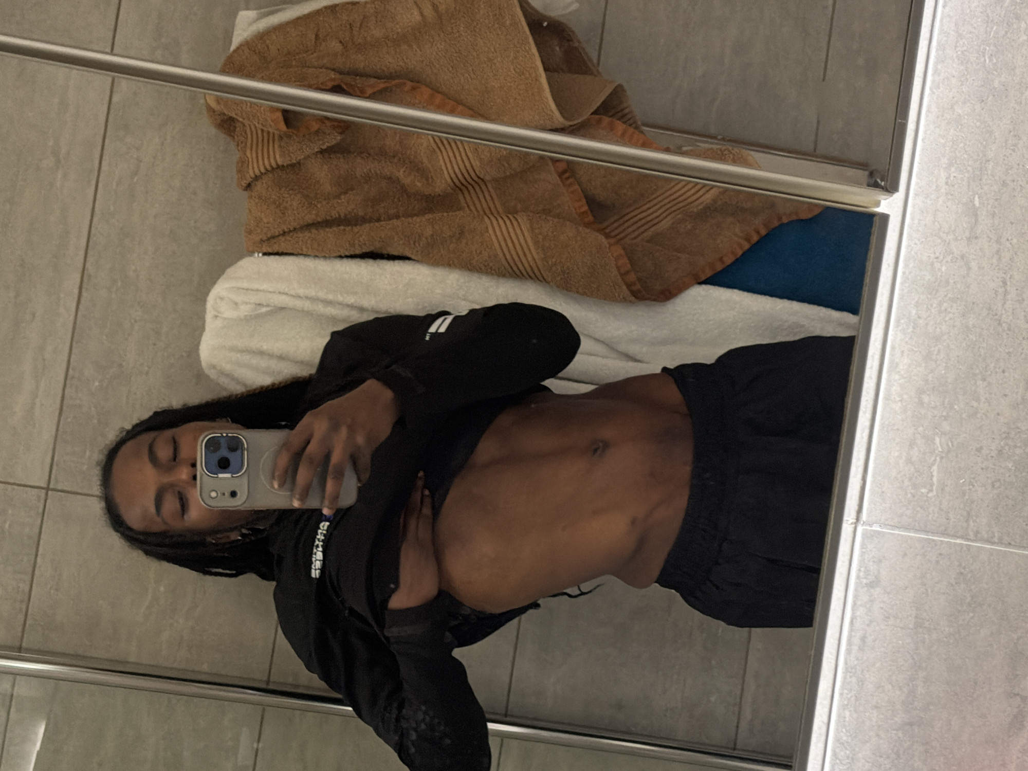 A mirror selfie, rotated 90 degrees clockwise, captures a person with dark skin and dreadlocks. Their eyes are closed as they hold a smartphone, an iPhone with a grey protective case and a triple-lens camera system, to take the photo. The person is wearing a black long-sleeved top, which is pulled up to reveal their midriff, showcasing visible and toned abdominal muscles. Black shorts or trousers are also part of their attire. White text, possibly "SUCCESS", is partially visible on the sleeve of their top.
The background features light grey rectangular tiles, indicating an indoor setting likely to be a bathroom or dressing room. In the reflection, behind the person, there's a stack of folded towels: a brown terry cloth towel is on top, with a white towel visible underneath, and a hint of blue fabric possibly below that. A horizontal silver metal bar, resembling a towel rack or part of a shower enclosure, extends across the frame. The overall impression is a personal moment captured in a reflective surface.