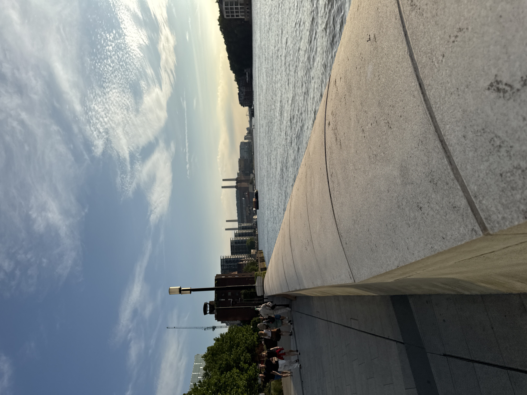 This is a vertical, medium-shot, eye-level photograph taken at sunset along the River Thames in London, United Kingdom. In the foreground, a curved, light-grey stone embankment wall dominates the right side of the frame, its surface textured with visible seams and small specks. To the left of the embankment is a paved walkway, and along this walkway, a group of six individuals are walking away from the camera. They are all dressed in casual clothing, with some carrying bags. Behind the walkway, there are trees, a tall building with dark windows, a dark brick structure, and industrial buildings with two prominent chimneys in the distance. The river, with gentle ripples on its surface, stretches across the middle and right portions of the image. The sky above is a blend of pale blue and golden hues, with wispy white clouds, indicating the setting sun. A lamppost with a modern, cylindrical light fixture stands tall on the embankment between the walkway and the river. The overall lighting suggests late afternoon or early evening.