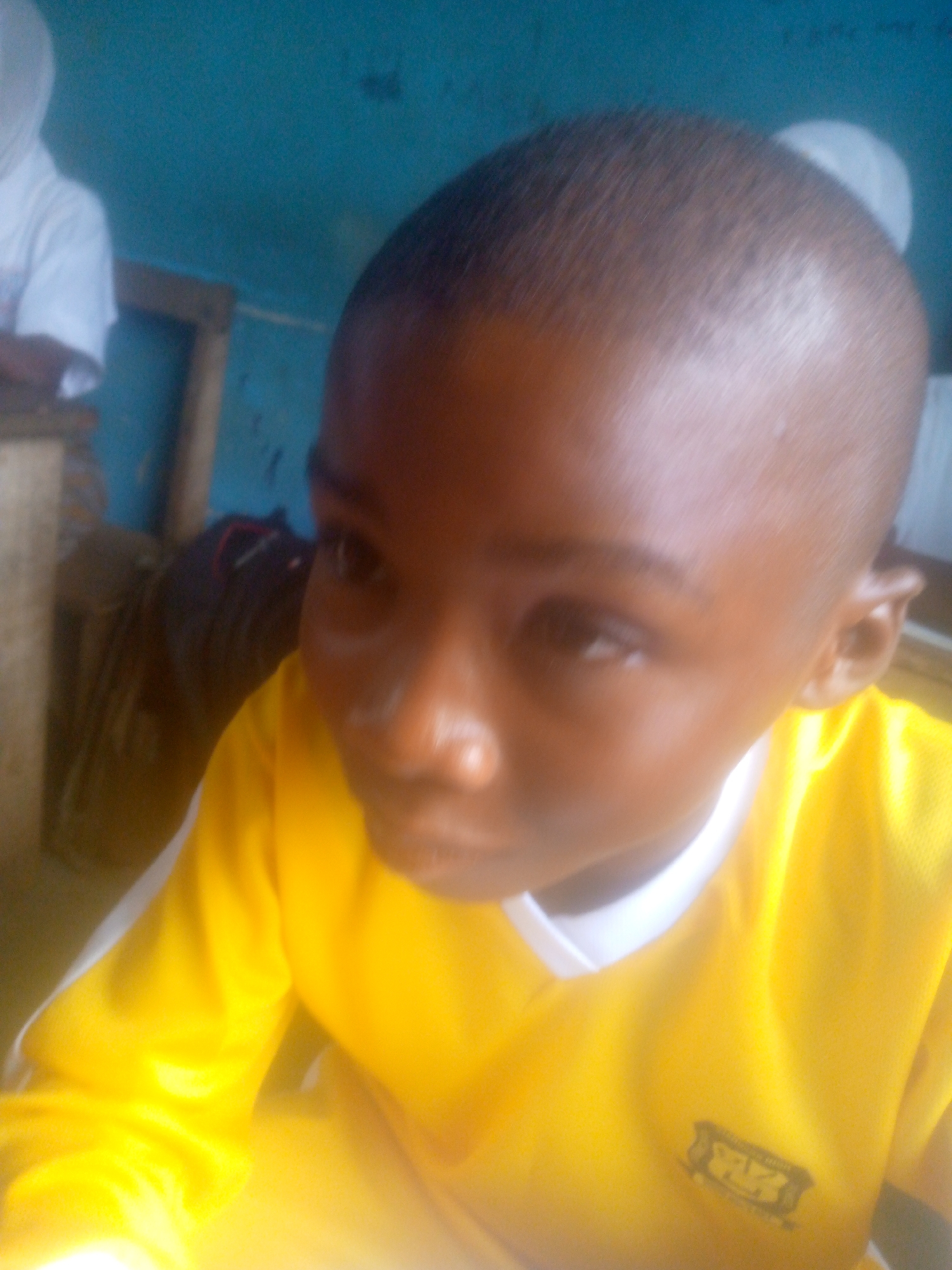 The image is a close-up shot of a young, dark-skinned child wearing a bright yellow shirt with a white collar. The child has a very short, close-cropped haircut. The child is looking slightly to the side with a focused or thoughtful expression. In the background, partially visible, is another person wearing a white shirt and seated at a wooden table. The background also includes a blue wall and what appears to be a dark-colored bag or object. The location is specified as Unknown, Unknown.