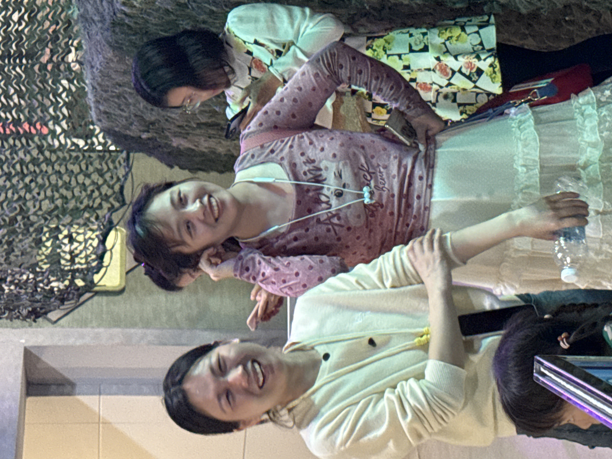 The image shows three women indoors. One woman is wearing a white sweater, and another woman is wearing a pink polka-dot shirt and a white tulle skirt. A third woman is wearing glasses and a white top with a floral pattern. The women appear to be interacting or posing for a photo. The photo was taken in 江北城街道, China.