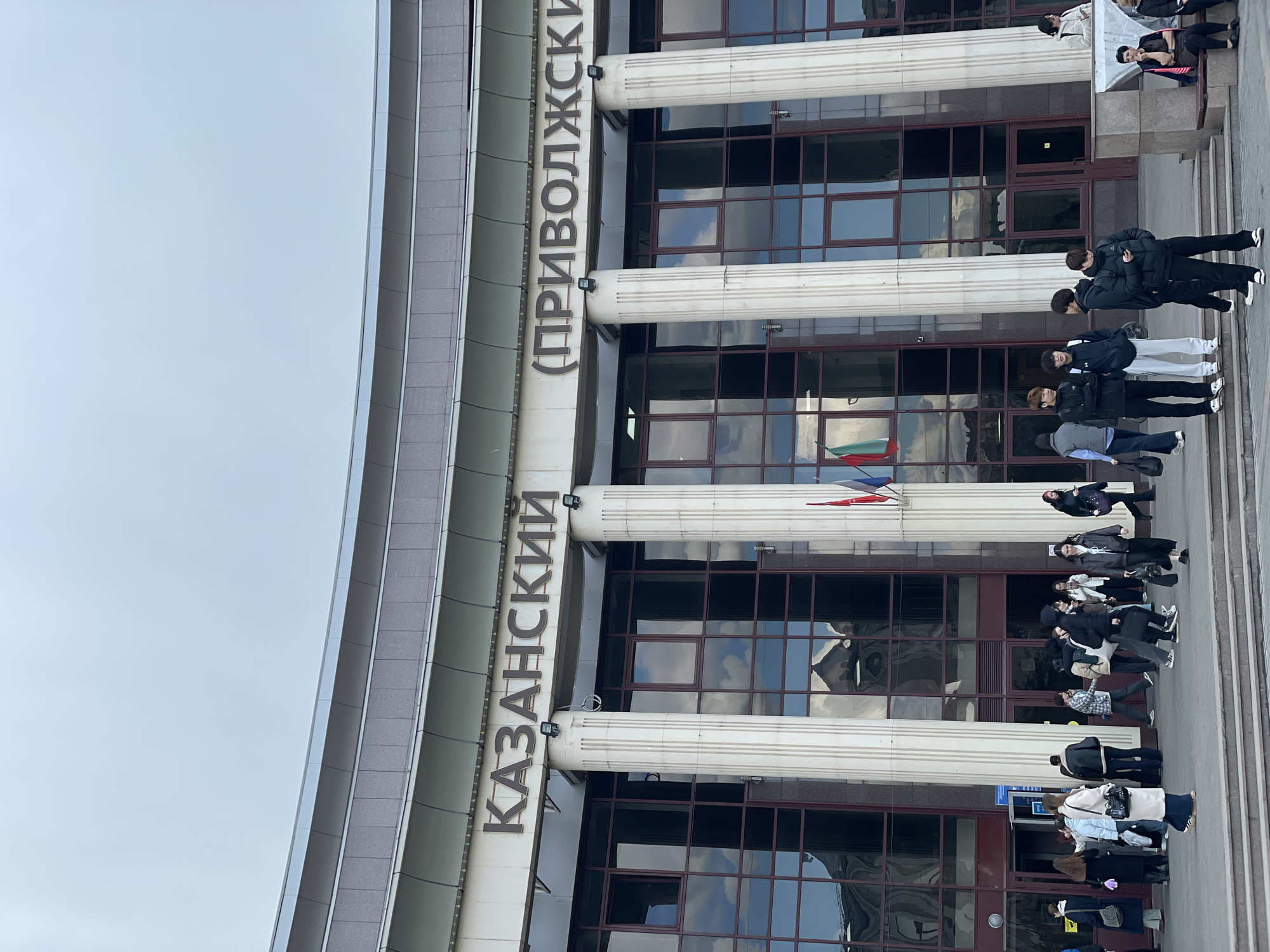 This vertically rotated image captures the facade of a large institutional building in Kazan, Russia, under an overcast, grey sky. The architecture features prominent classical elements, including several tall, fluted columns of a light color, set against a dark-windowed mid-section and a grey-paneled upper structure. The windows reflect the cloudy sky and hints of the surroundings.
Large, dark Cyrillic text is displayed horizontally across the building's facade, reading "КАЗАНСКИЙ (ПРИВОЛЖСКИЙ)". This translates to "Kazan (Volga Region)", clearly indicating the building's location and its likely association with a significant institution in Kazan, possibly Kazan Federal University. To the left of the building's entrance, a flag with horizontal stripes, appearing to be the flag of Tatarstan (green, white, red), is partially visible.
A moderate crowd of people, mostly young adults, are gathered on the wide pavement and steps leading to the building's entrance. They are dressed in casual outerwear, such as jackets and coats, suggesting cool weather. Many are standing in small groups, conversing or waiting, while others appear to be walking to or from the building. On the right side of the frame, some individuals are seen on a raised platform or ledge. The scene depicts a typical day with people congregating and moving about in front of a significant public edifice.