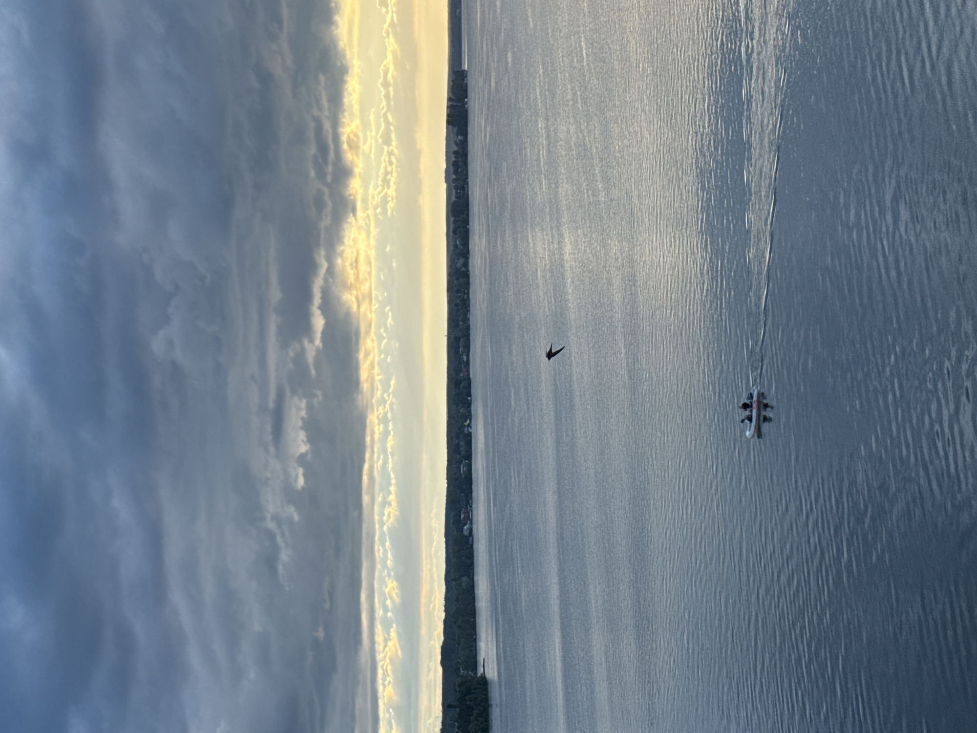 The image shows a wide, calm body of water, likely a lake, with a distant shoreline lined with trees and scattered buildings. The sky is filled with dramatic, dark clouds, with a bright band of golden light peeking through near the horizon, suggesting either sunrise or sunset. In the foreground, a small, white inflatable boat with two figures in it is being paddled across the water, leaving a faint wake. A single bird is in flight in the middle distance, its silhouette sharply defined against the sky. The overall mood is serene yet a little moody due to the overcast sky. The location is the Votkinsk Reservoir in Votkinsk, Russia.