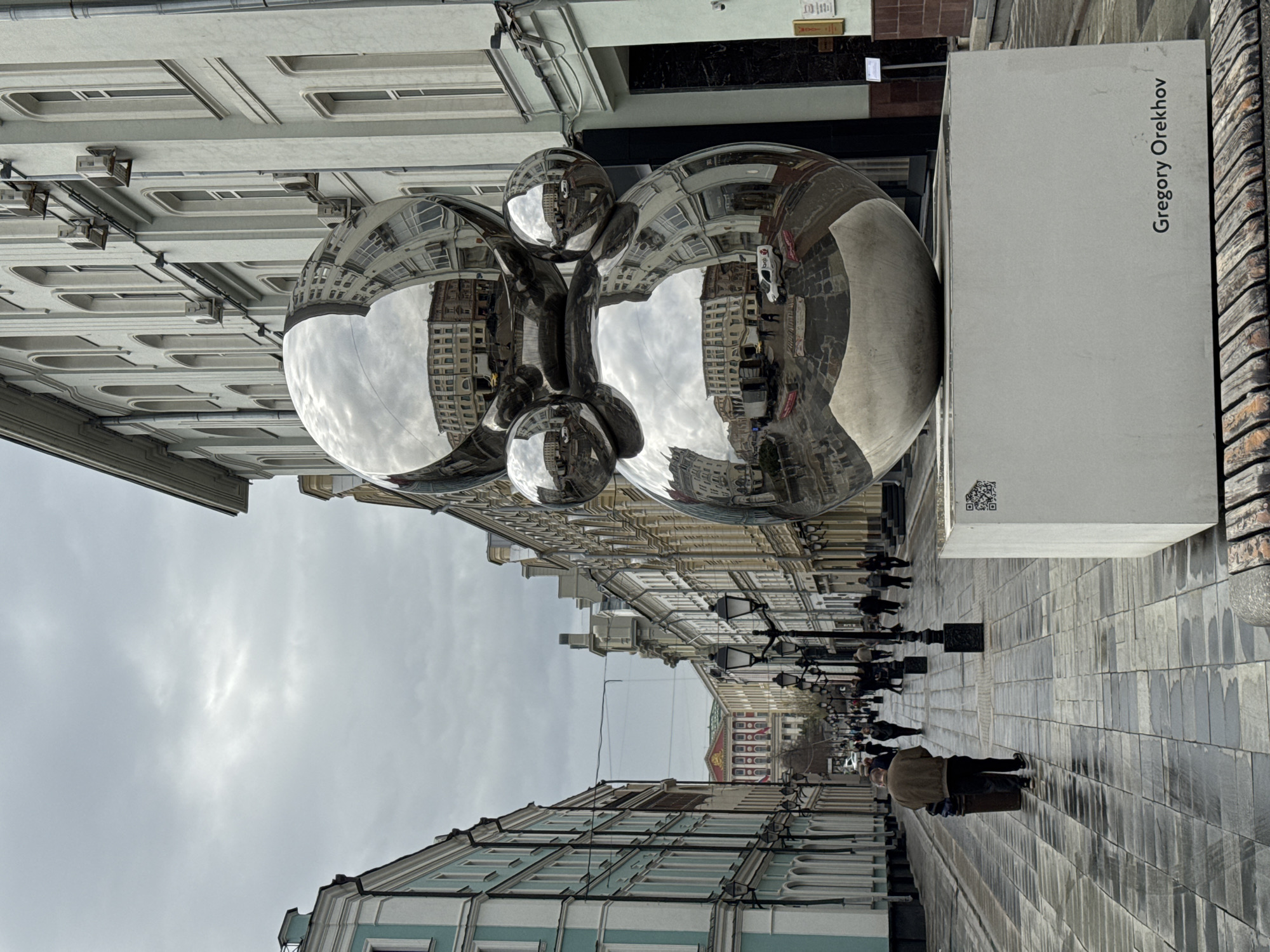 The image captures an urban pedestrian street, identified as being in Moscow, Russia, featuring a prominent contemporary sculpture. The central subject is a large, highly reflective, chrome-plated sculpture composed of multiple connected spherical and elongated forms, resembling inflated balloons or abstract organic shapes. This sculpture stands on a white rectangular pedestal, which clearly displays the name "Gregory Orekhov" in dark lettering, along with a QR code.
The polished surface of the sculpture acts as a mirror, vividly reflecting the surrounding buildings, the overcast sky, and the streetscape, creating distorted yet recognizable images within its surface.
The street itself is paved with light-colored rectangular blocks, some appearing subtly damp or reflective. Buildings with classical architectural features and multiple stories line both sides of the thoroughfare. Those in the foreground on the left are painted in light blue and white hues. Streetlights with multiple lamps are spaced along the pedestrian zone.
Several people are visible, casually walking along the street in various directions, with some figures partially captured in the sculpture's reflections. The sky is overcast and bright, indicating a daytime setting with diffused light. The overall scene portrays a typical urban environment, enhanced by the striking modern art installation that interacts dynamically with its historical surroundings through its reflective properties.