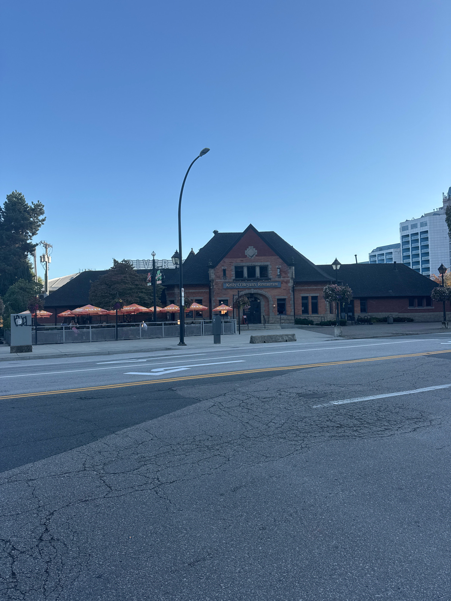 The image shows the exterior of Kelly O'Bryan's Restaurant, a building with red brick construction and a dark shingled roof. The restaurant has a sign above its entrance. Orange umbrellas are set up on an outdoor patio area adjacent to the building. In front of the restaurant, there is a sidewalk and a street with visible lane markings and some cracking in the asphalt. Tall trees are present on the left side of the image, and modern buildings are visible in the background to the right. The sky is clear and blue. The location is identified as New Westminster, Canada, and specifically the building is located at 800 Columbia Street.