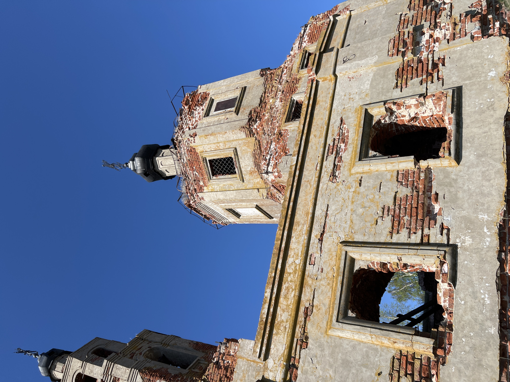 The image shows a low-angle view of a dilapidated building in Шуран, Russia, against a clear blue sky. The building appears to be a church or religious structure, judging by the presence of onion-shaped domes with crosses on top. The structure is made of brick, covered with a layer of plaster or concrete that is peeling and crumbling, revealing the brick beneath. The walls have several window openings, some of which are damaged and show signs of decay, with remnants of wooden frames visible. The overall condition of the building indicates significant neglect and disrepair.