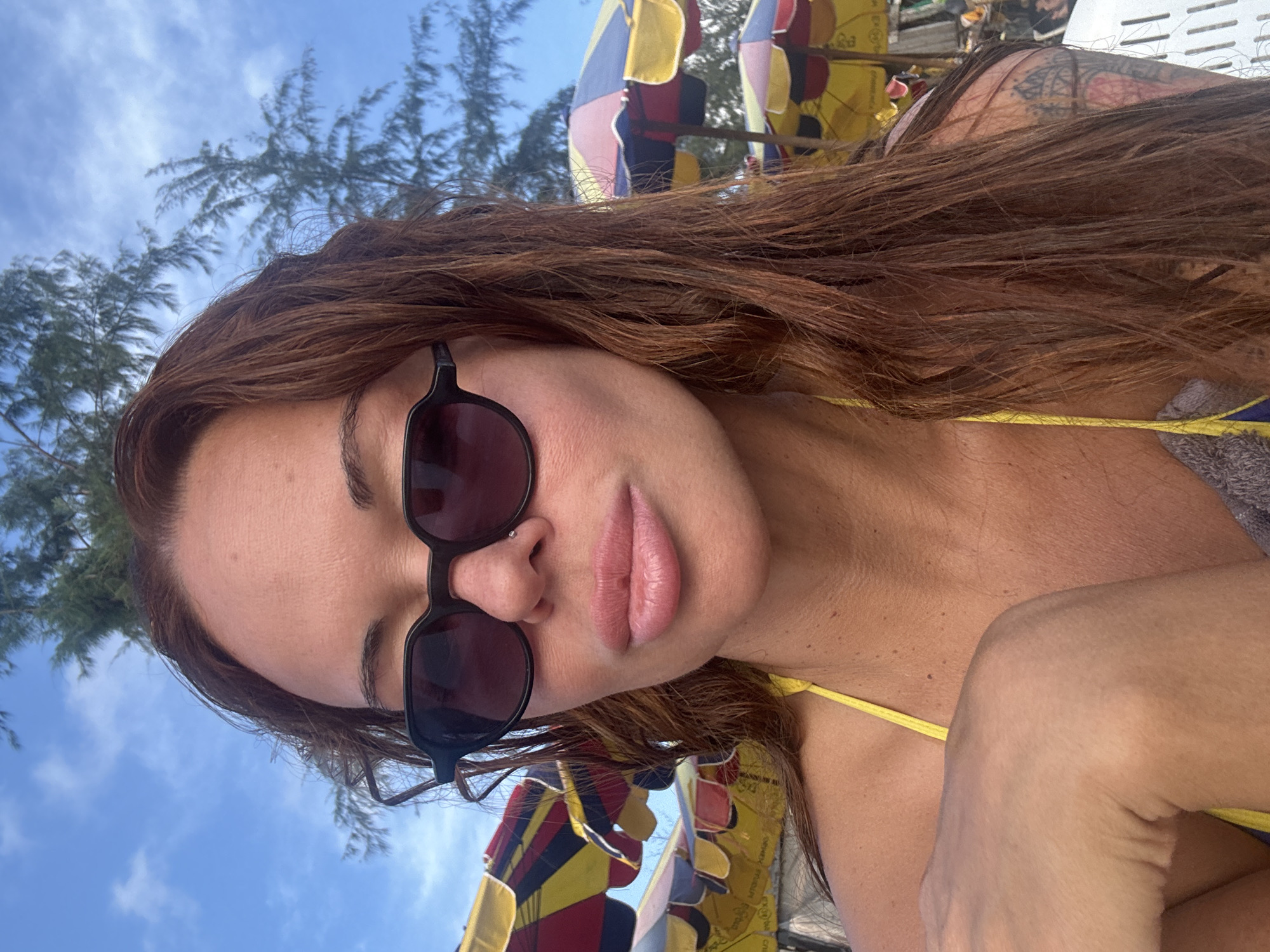 In a close-up, outdoor selfie taken at a beach in Choeng Thale, Thailand, a woman with long, reddish-brown hair and sunglasses is the primary subject. She is wearing a yellow bikini top. The background reveals a bright blue sky with scattered clouds and trees, as well as a row of colorful beach umbrellas. The woman's face is turned towards the camera, with her mouth slightly open and her lips appearing full. A subtle nose piercing is visible. Her right arm is bent, with her hand resting on her chest. The lighting suggests it is daytime, likely during sunny weather.
