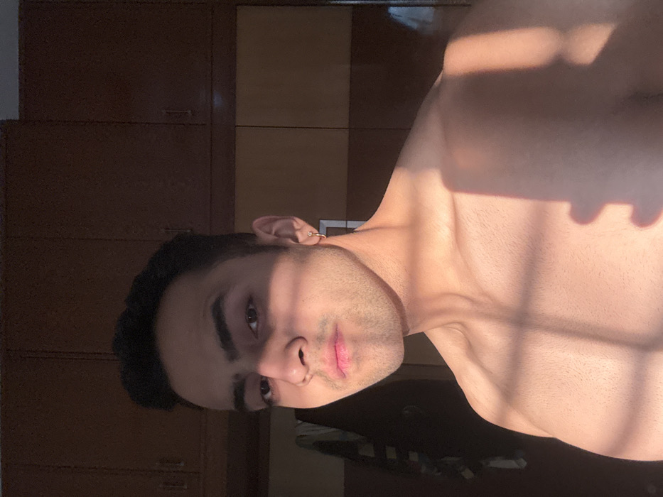 A shirtless man faces the camera, sunlight casting shadows across his face and chest. He has dark hair and eyes, with stubble on his face and a ring earring. He is positioned in front of dark wooden cabinets. The photo is turned sideways.