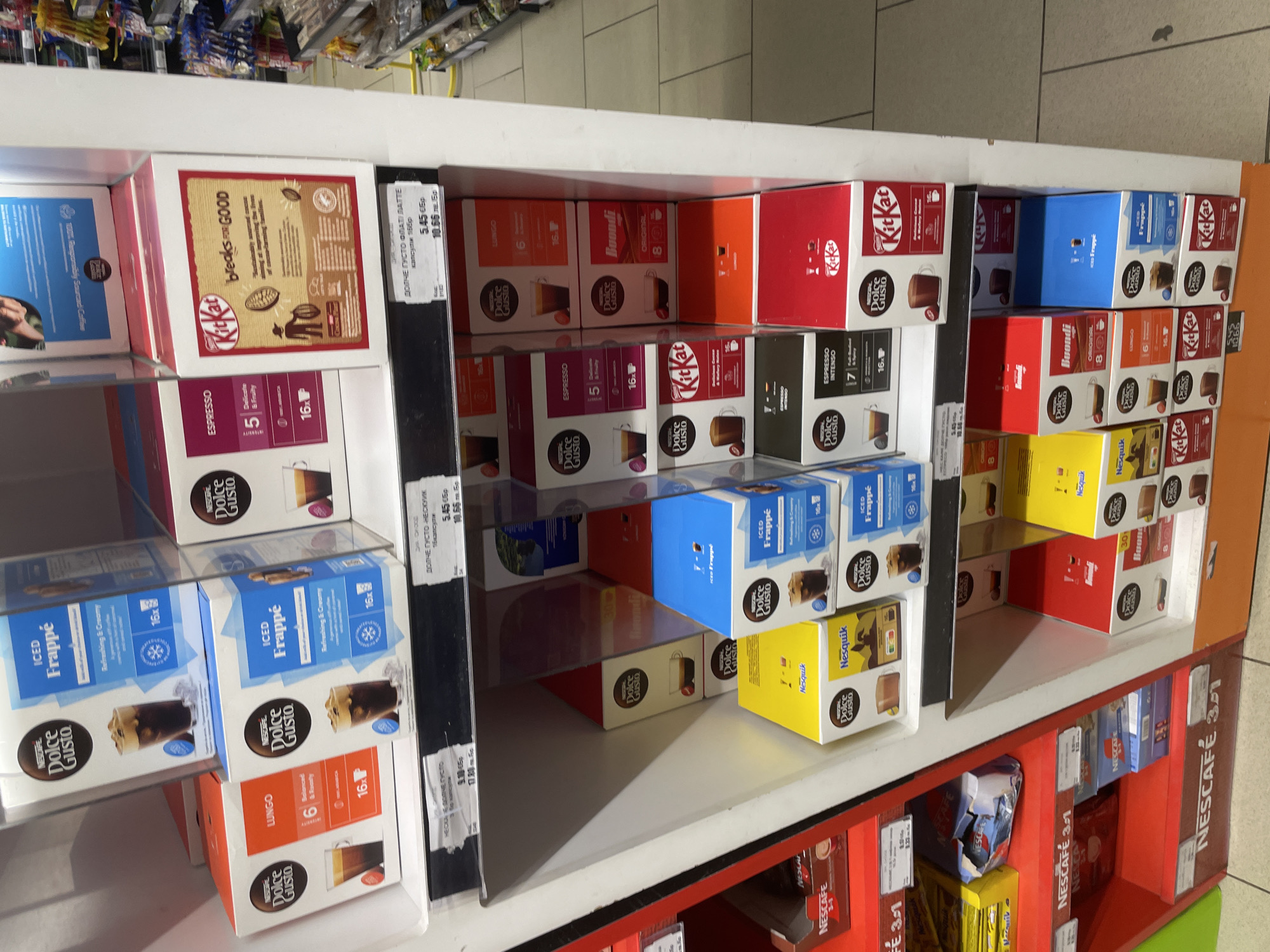 The image captures a retail shelf in Ihtiman, Bulgaria, displaying a variety of Nescafe Dolce Gusto coffee capsules and related beverage products. The scene is an interior shot, likely within a supermarket or grocery store, characterized by artificial lighting.
The main subjects are numerous colorful boxes of coffee pods and hot chocolate mixes, neatly organized on white shelves. Prominent brands include Nescafe Dolce Gusto, KitKat, Buondi, and Nesquik. Various product types and flavors are visible, such as Espresso, Lungo, Espresso Intenso, and Iced Frappé, along with Nesquik for children. Many boxes indicate they contain "16x" capsules, while some Buondi boxes specify "8x". Descriptive text like "Balanced & Roasty," "Refreshing & Creamy," and "Delicate & Fruity" is also present, often alongside illustrations of prepared drinks. One KitKat box features the slogan "KitKat breaks for GOOD."
Price tags are attached to the shelf edges. These tags display prices in Bulgarian Leva (лв. Бр) and, in some instances, also in Euros (€Бр). Visible text on the tags includes "ДОЛЧЕ ГУСТО ФЛАТ ЛАТТЕ капсули 16бр" (Dolce Gusto Flat Latte capsules 16pcs) and "ДОЛЧЕ ГУСТО НЕСКУИК капсули 16бр" (Dolce Gusto Nesquik capsules 16pcs), both priced at 5.45 €Бр / 10.66 лв. Бр. Another partially visible tag mentions "НЕСКАФЕ" with a price of 9.10 €Бр / 17.80 лв. Бр. On a lower shelf, boxes of "NESCAFE 3-1" (Nescafe 3-in-1) are also visible.
The shelves show some empty spaces, suggesting product turnover or stock replenishment in progress. The image is rotated 90 degrees clockwise. No people are visible, and no specific activity is taking place beyond the static display of goods for sale.