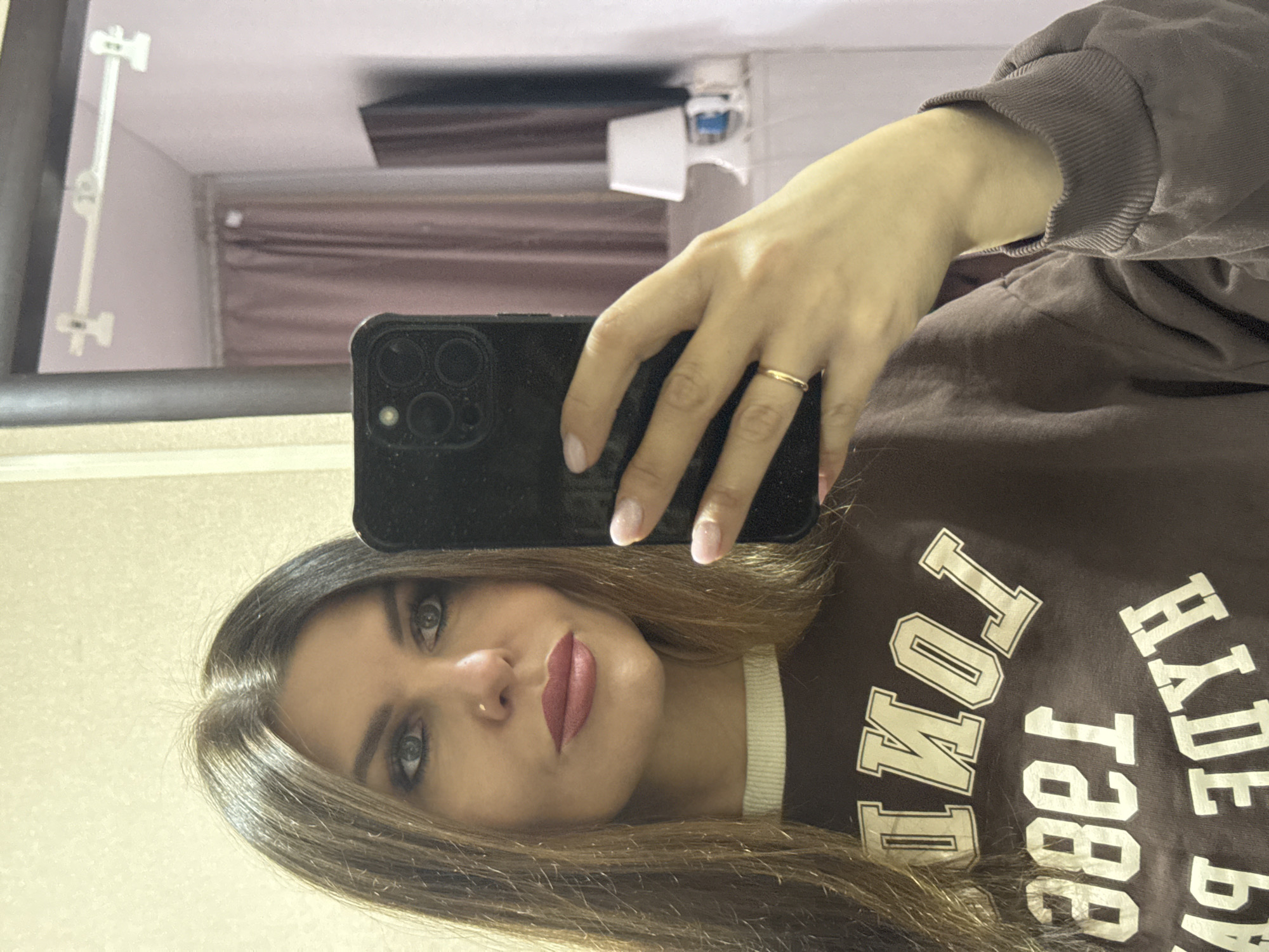 A young woman with light brown hair and gray eyes takes a selfie in a mirror. She wears a brown sweatshirt with "Hyde Park 1866 London" printed on it. She holds a black smartphone with her right hand, a simple gold ring on her finger. The reflection in the mirror shows a room with pink curtains and a clothes-drying rack attached to the wall. There is no evidence of the location being Chaikovsky, Russia.