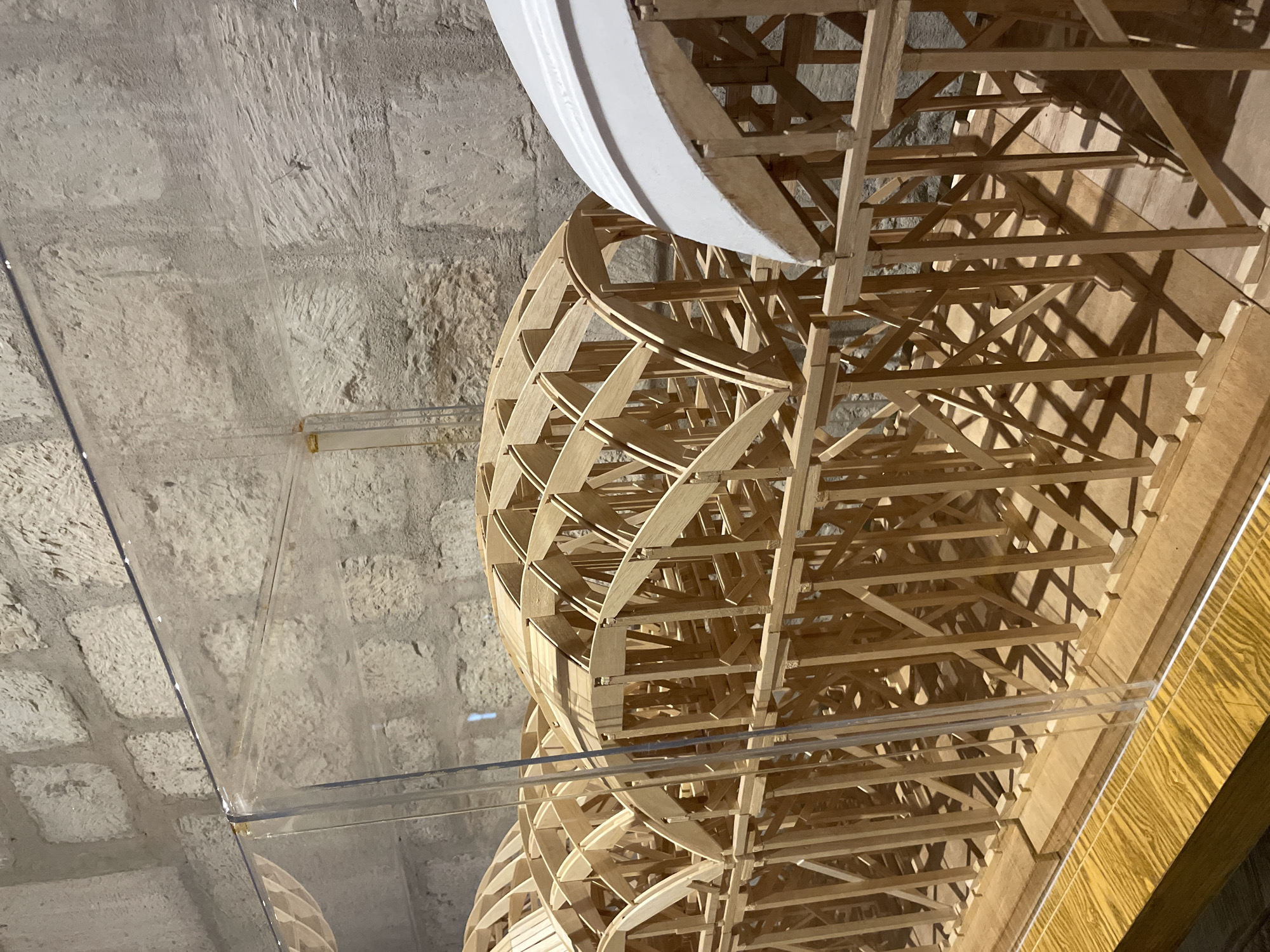 This image depicts a wooden lattice structure, likely a model or artistic installation, enclosed within a clear acrylic display case. The wooden structure is complex, featuring curved elements and intricate intersections. The background includes a rough, textured wall, possibly stone or plaster, adding to the aged or historical atmosphere. The presence of the display case suggests the structure is being preserved or presented for public viewing, and the surrounding architectural details suggest the location is indoors, possibly in a museum or gallery. While I do not have the ability to verify its location in Oaxaca City, Mexico, the architectural details provide clues about the possible geographical setting.
