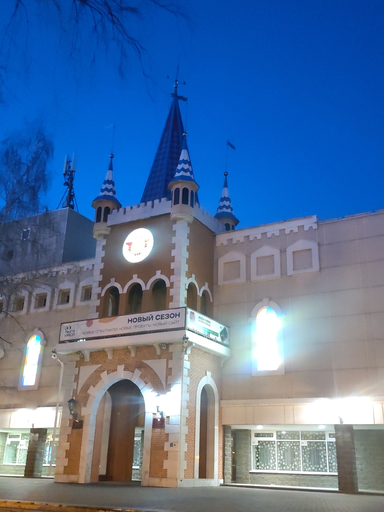 The image displays the illuminated facade of a puppet theater in Izhevsk, Russia, at dusk or early night. The building is designed to resemble a fairytale castle, featuring light brown brickwork, white trim, and multiple turrets with distinctive blue and white patterned conical roofs. A prominent central spire, topped with a weather vane, rises above the main structure.
A large, circular, glowing clock face is visible above the central arched entrance. Below this, a long banner announces "НОВЫЙ СЕЗОН" (NEW SEASON) and "НОВЫЕ СПЕКТАКЛИ, НОВЫЕ ПРОЕКТЫ, НОВЫЙ САЙТ" (NEW PERFORMANCES, NEW PROJECTS, NEW WEBSITE), indicating upcoming events at the venue. Other visible text includes "ТЕАТР КУКОЛ" (PUPPET THEATER) and "ВЕДИ СЕБЯ Культурно" (BEHAVE Culturally).
The sky is a deep, clear blue, indicative of the twilight hours. Bare tree branches are visible against the sky on the left side of the frame. The building is brightly lit by numerous artificial lights, causing significant lens flares around windows and other light sources. Communication antennas are visible on a flat roof section to the left of the main castle-like structure. The scene is devoid of people, focusing solely on the architectural details and the evening atmosphere of the theater.