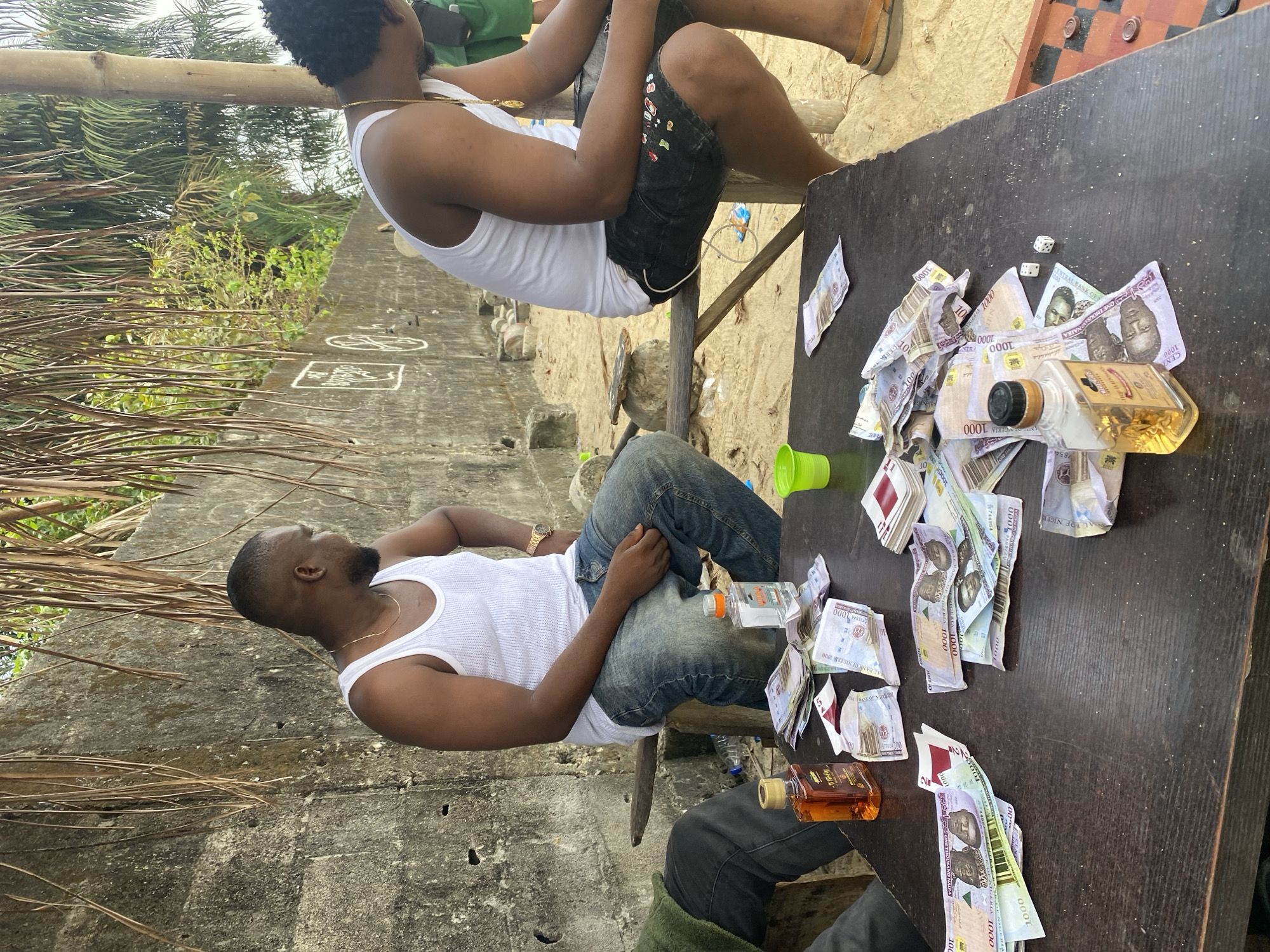 Here is a detailed description of the image:
**Scene Overview:** The image captures a casual, relaxed scene outdoors. There are at least two people, money, and alcoholic beverages.
**People:**
* Two men are visible. One man wears a white tank top and blue jeans and is sitting down. A second man, also wearing a white tank top and dark shorts, sits above, likely on some kind of makeshift seating. A third person is cropped off in the bottom right corner, but their black jeans and green coat are visible.
**Objects:**
* A black table is covered with a large amount of Nigerian Naira (paper money).
* At least two bottles, one of which has brown liquid, are placed on the table among the money. One bottle contains clear liquid.
* A green plastic cup is also on the table.
* A checkered board is visible on the upper right of the table.
* The wall in the background is light grey, and has some markings on the upper left, including two squares and two circles.
**Environment:**
* The scene appears to be outdoors, possibly in a backyard or open space. The ground consists of sand.
* The environment seems casual and informal, implying a relaxed setting.