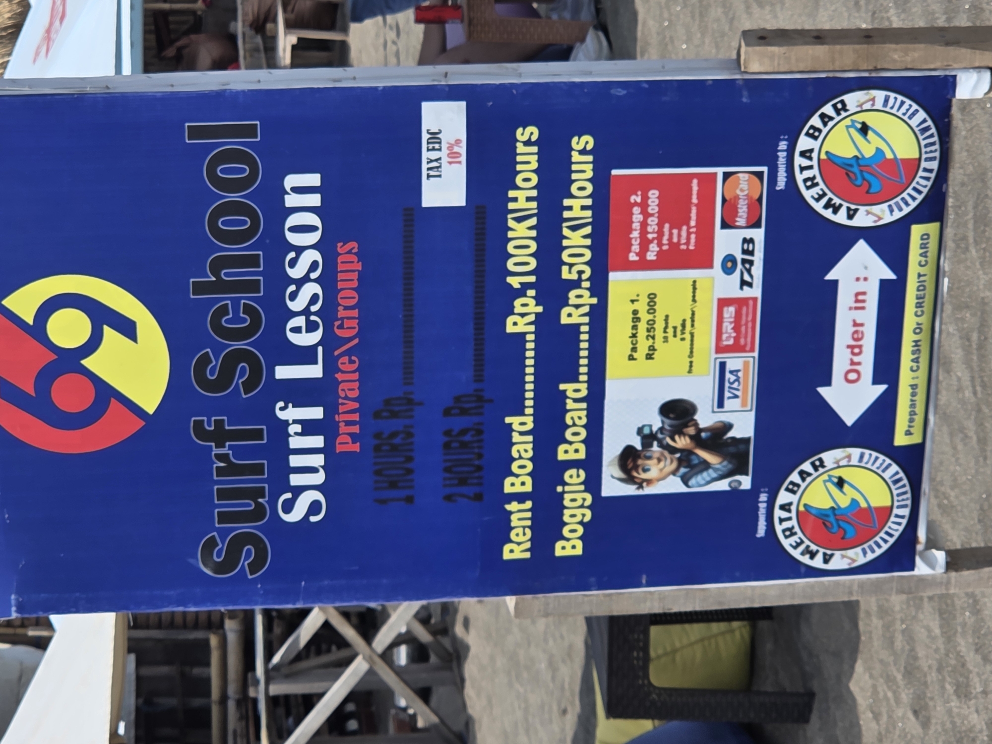The image shows a sign for a surf school, likely located in North Kuta, Indonesia. The sign is blue with yellow and red lettering and displays information about surf lessons, board rentals, and packages. It lists the prices for 1 and 2-hour lessons, board rentals, and bogie board rentals. Two packages are listed: Package 1 costs Rp.250,000 and includes 10 photos and free coconut water, while Package 2 costs Rp.150,000 and includes 3 videos and free water. The sign also indicates that a 10% tax (EDC) is applicable. It features logos of credit card providers and says "Supported by: AMERTA BAR". An arrow pointing upwards reads "Order in:". The bottom right corner reads "Prepared: CASH or CREDIT CARD". A cartoon image of a person with a camera is also visible. The background of the image shows a beach setting with people and structures.