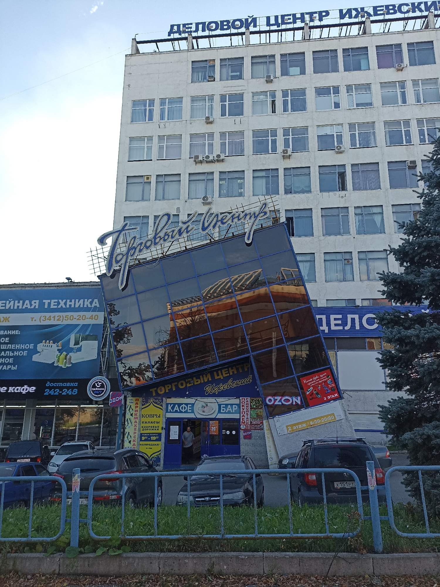 This is an outdoor, eye-level, full shot photograph of the "Delovoy Tsentr" (Business Center) building in Izhevsk, Russia. The building is a multi-story, light-colored structure with many windows. A large, angled glass-fronted section protrudes from the building, displaying signage for "Torgovyy Tsentr" (Shopping Center). The glass reflects the sky and parts of the building.
Several signs are visible: "DELOVOY Tsentr IZHEVSKIY" at the top of the building, and other signs for various shops and services, including "MAI" and "CHAYKA" (likely related to sewing machines and a 24/7 delivery service), and "OZON" (an online retailer). There is also a sign indicating "2 ETAZH" (2nd Floor) with operating hours.
In front of the building, parked cars are visible on a paved area and on grass behind a blue metal fence. A man wearing a grey t-shirt is standing in an open doorway of one of the shops. The scene appears to be in an urban commercial area. The lighting suggests it is daytime, with a clear sky.