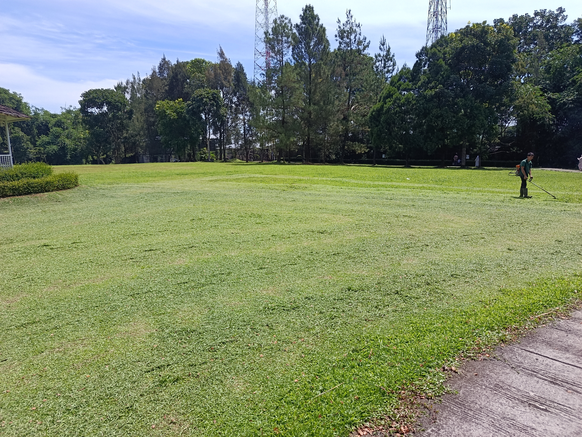 The image depicts a large, well-maintained lawn in Bandar Baru, Indonesia. A man in a green shirt and dark pants is operating a string trimmer on the lawn, indicating he is likely a groundskeeper or gardener. The lawn is surrounded by lush green trees, with tall communication towers visible in the background. The sky is a clear blue with scattered white clouds. A paved walkway runs along the right side of the image, bordering the lawn. The overall scene suggests a park, golf course, or similar public green space within Bandar Baru.