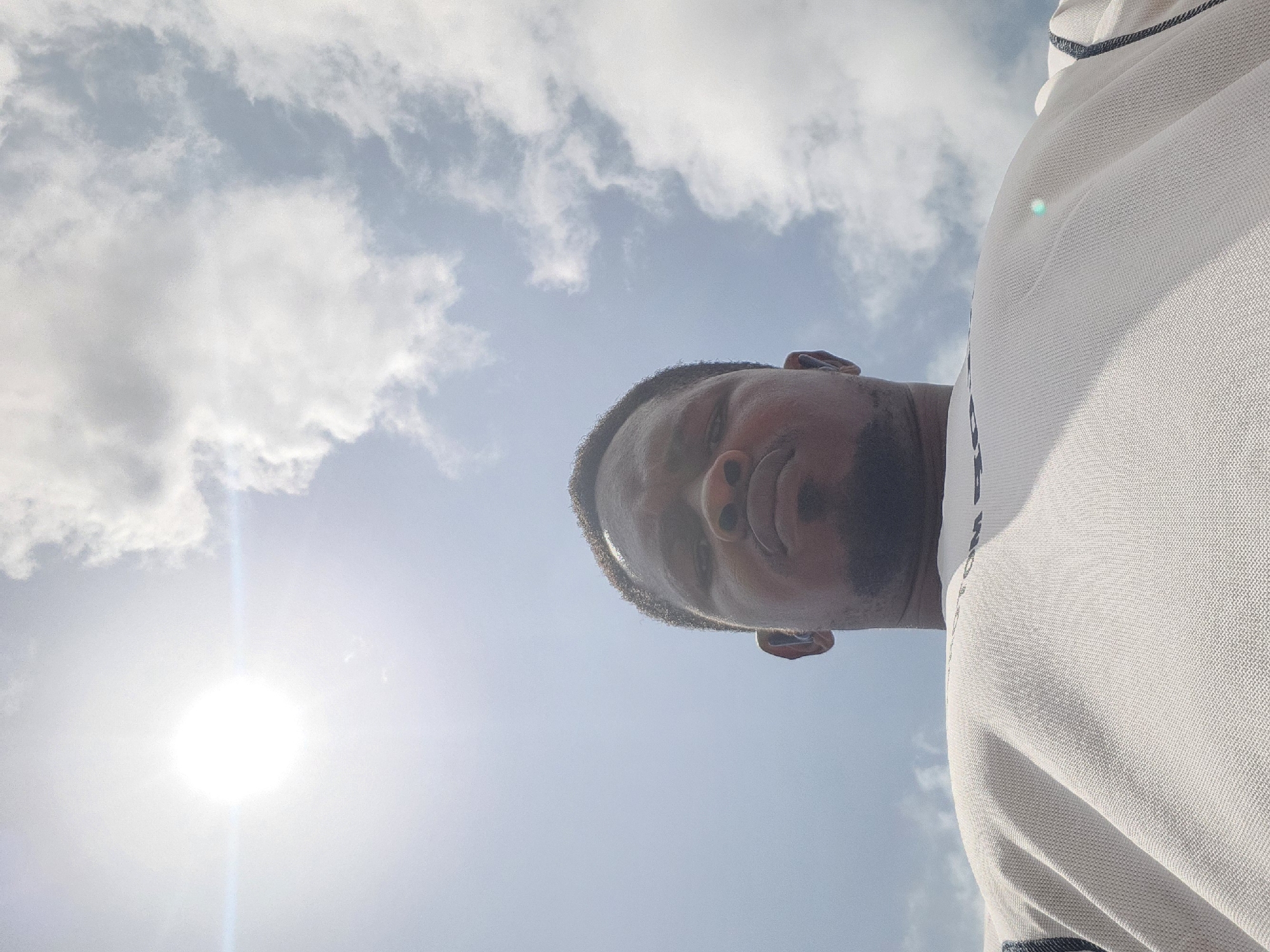 A low-angle, eye-level shot focuses on the face of a man with dark skin and short hair, looking upwards at the sky. He is wearing a white shirt. The sun is bright and high in the sky, creating a lens flare that extends horizontally across the image. Clouds are visible in the sky. The image is taken outdoors in daylight.