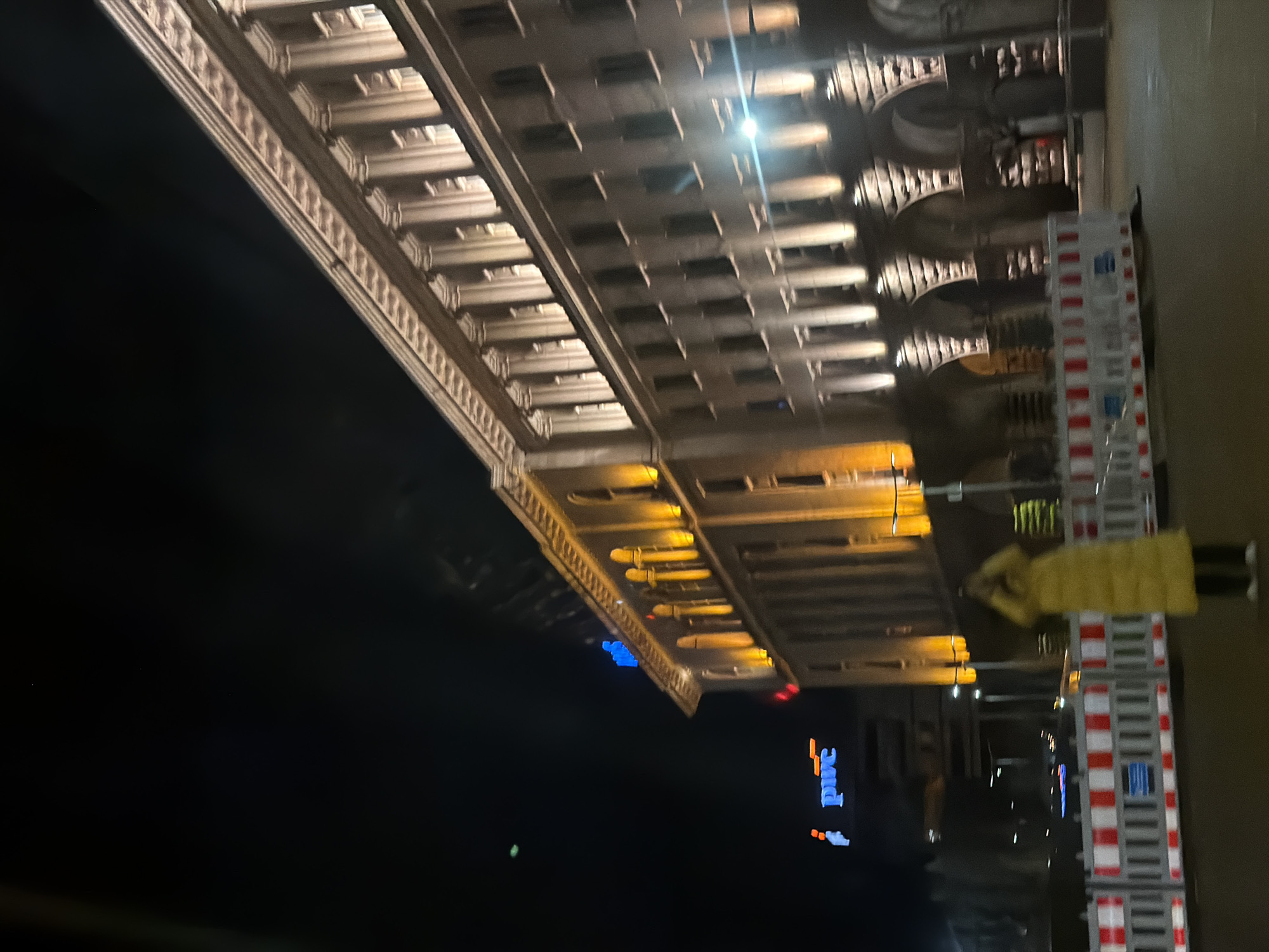 The image shows the illuminated facade of a building at night. The architectural style suggests it could be located in a European city. There is also a construction worker wearing a yellow vest and a metal scaffolding structure. A sign that reads "PMC" with illuminated blue letters can also be seen. Based on the building style and sign, the image is likely of the facade of a building in Sofia, Bulgaria, with some street repair work taking place.