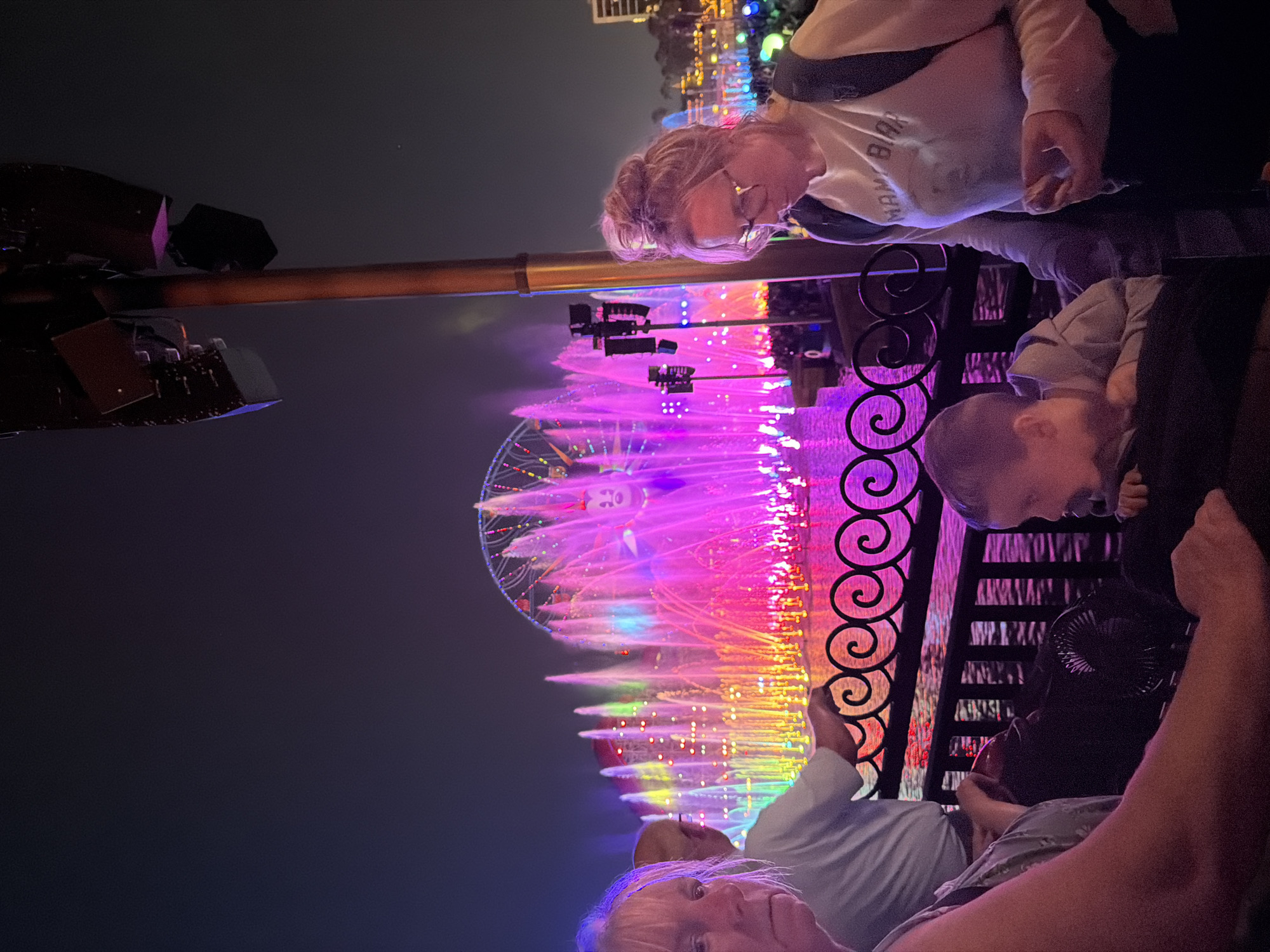 The image depicts a nighttime view of the World of Color show at Disney California Adventure Park in Anaheim, USA. Colorful fountains and lights illuminate the scene, with the Pixar Pal-A-Round ferris wheel visible in the background. A family is observing the show from behind a black metal fence. An adult with glasses and a white shirt is standing with a young child beside them, both looking towards the show. Another adult is seated with another young child behind the fence.