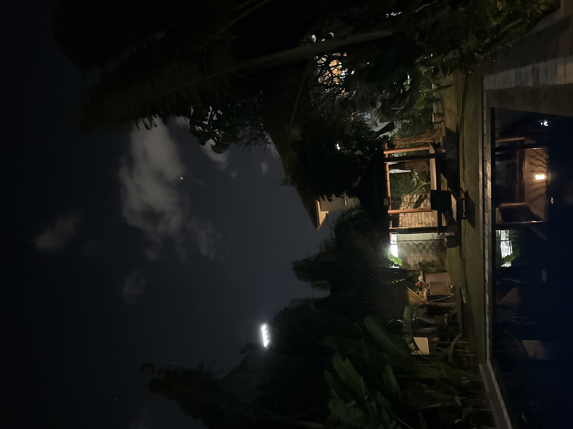 A nighttime outdoor scene shows a swimming pool in the foreground with a reflection of a gazebo and a lightbulb. The gazebo is situated on a grassy area behind the pool, with lounge chairs visible beneath it. Beyond the gazebo, a building with lights on can be seen, partially obscured by trees. The sky above is dark with some clouds and a few visible stars. The scene appears to be a private residence or resort, possibly in the city of Denpasar, Indonesia, given the lush tropical foliage. The lighting is dim, primarily from artificial sources within the scene and a bright rectangular light in the distance.