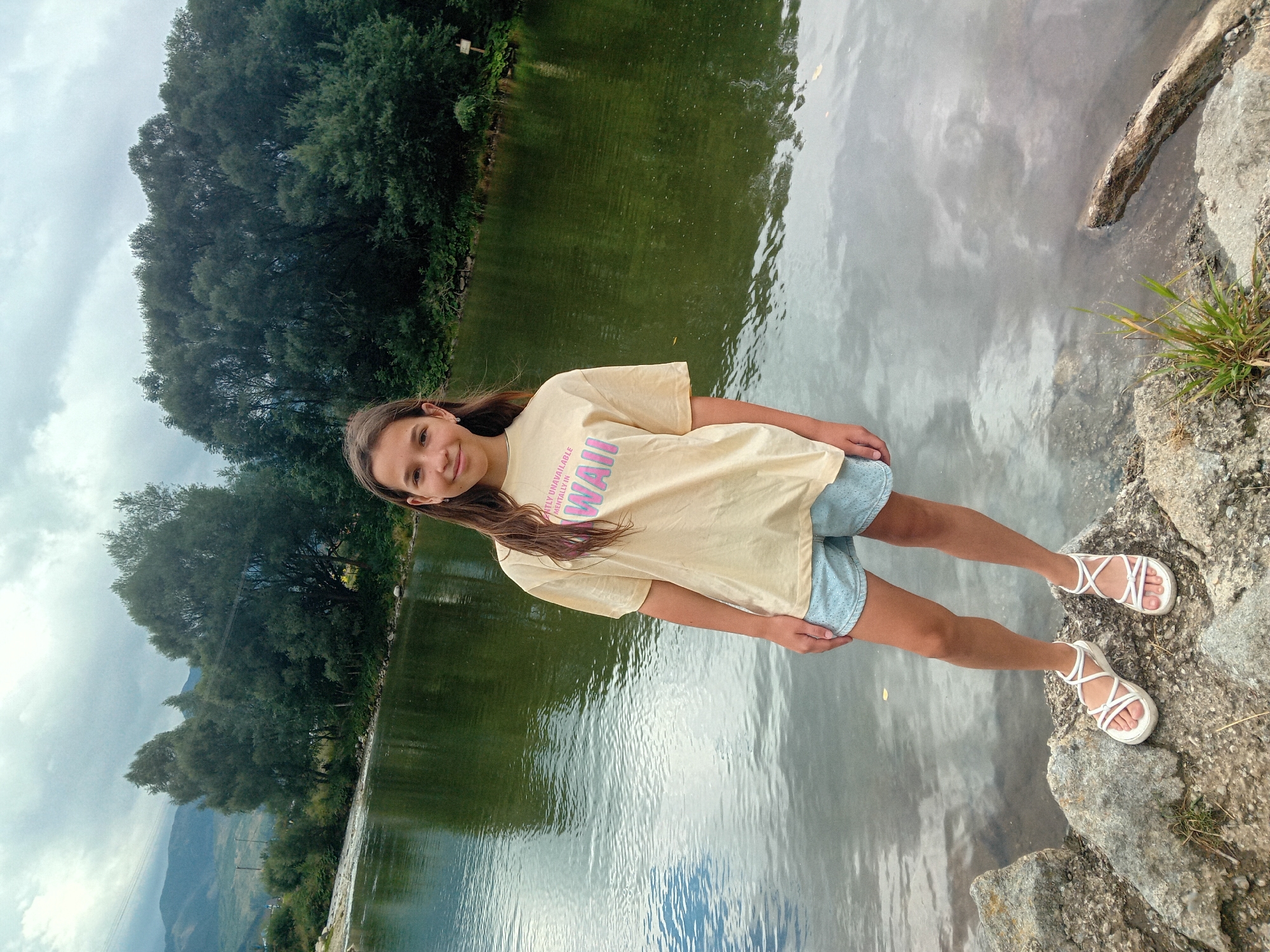 A young girl with long brown hair is standing by a lake, looking at the camera with a smile. She is wearing a light yellow t-shirt with pink and purple text that reads "UNAVAILABLE MENTALLY IN HAWAII" and light blue shorts with white polka dots. She is also wearing white strappy sandals. The girl is standing on a rocky shore, with a dense forest on the left side of the image and the lake reflecting the cloudy sky on the right. In the background, mountains can be seen through the trees. The time of day appears to be afternoon, and the weather is overcast. The scene is set in Habovka, Slovakia.