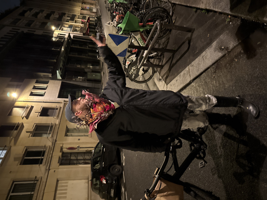 A man wearing a floral mask and a baseball cap stands next to a bicycle on a city street at night. He is wearing a dark jacket and boots and raises one arm in the air. Behind him are parked bicycles and a building with lit windows. A black car is parked nearby on the street.