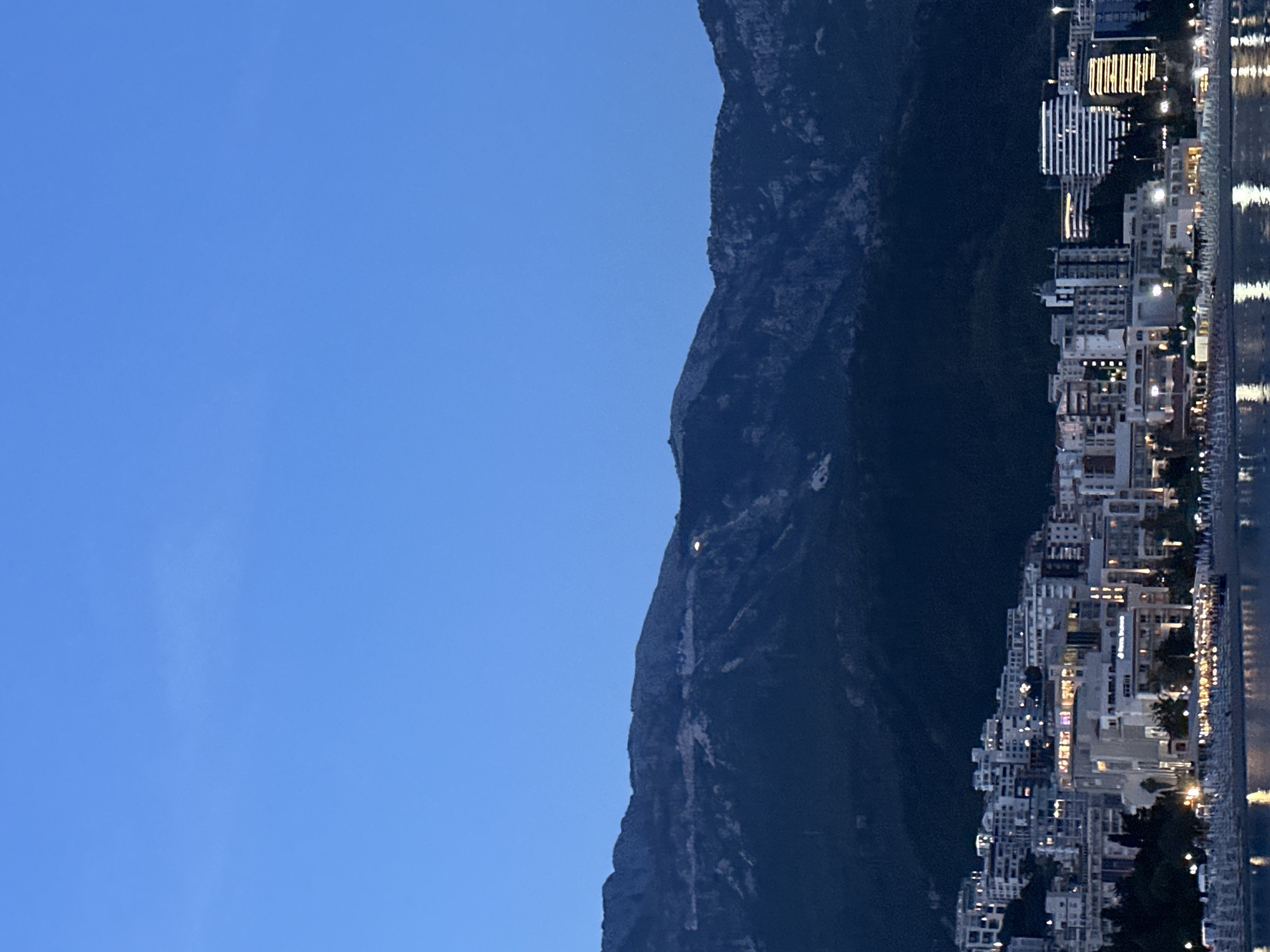The image depicts a night view of Budva, Montenegro, with a mountain range and cityscape visible. The sky is a dark blue, and the mountain is silhouetted against it. The city is illuminated with lights from buildings and street lamps. The scene includes the illuminated coastline. The image appears to be taken at dusk or dawn. The entire image is rotated 90 degrees clockwise.