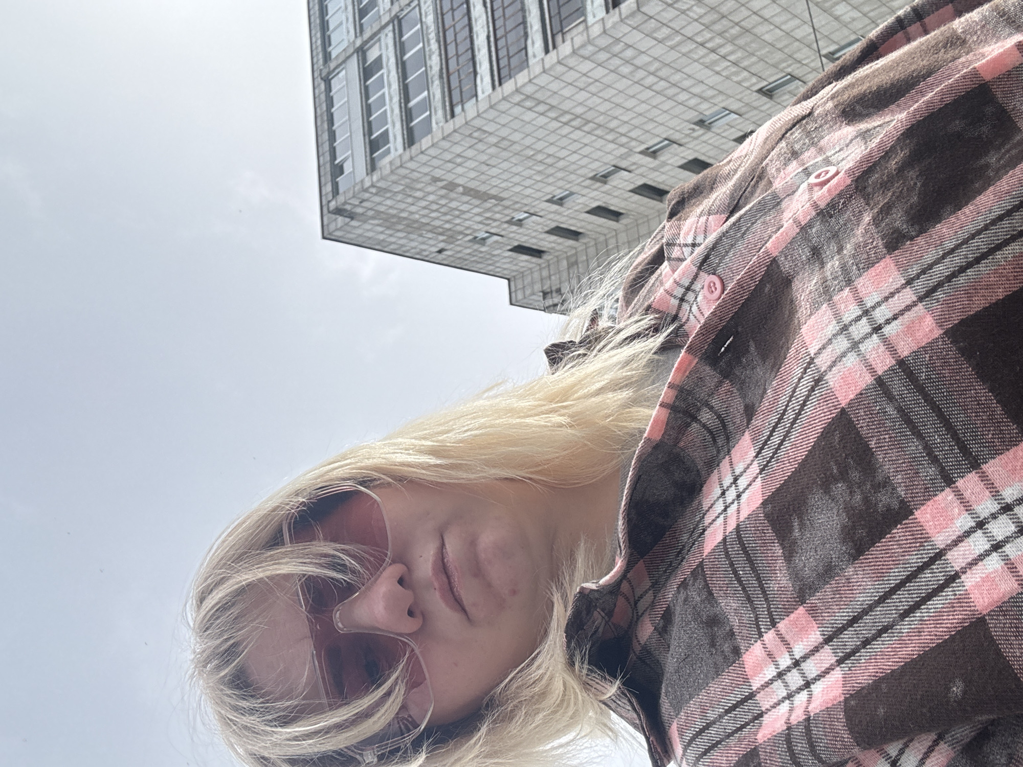 A low-angle, close-up shot captures a fair-skinned individual with blonde hair wearing pink-tinted sunglasses and a brown and pink plaid shirt. The person is looking downward. Behind the individual, a tall, tiled building is visible, suggesting an urban setting. Based on the location provided, this is most likely in Chaikovsky, Russia.