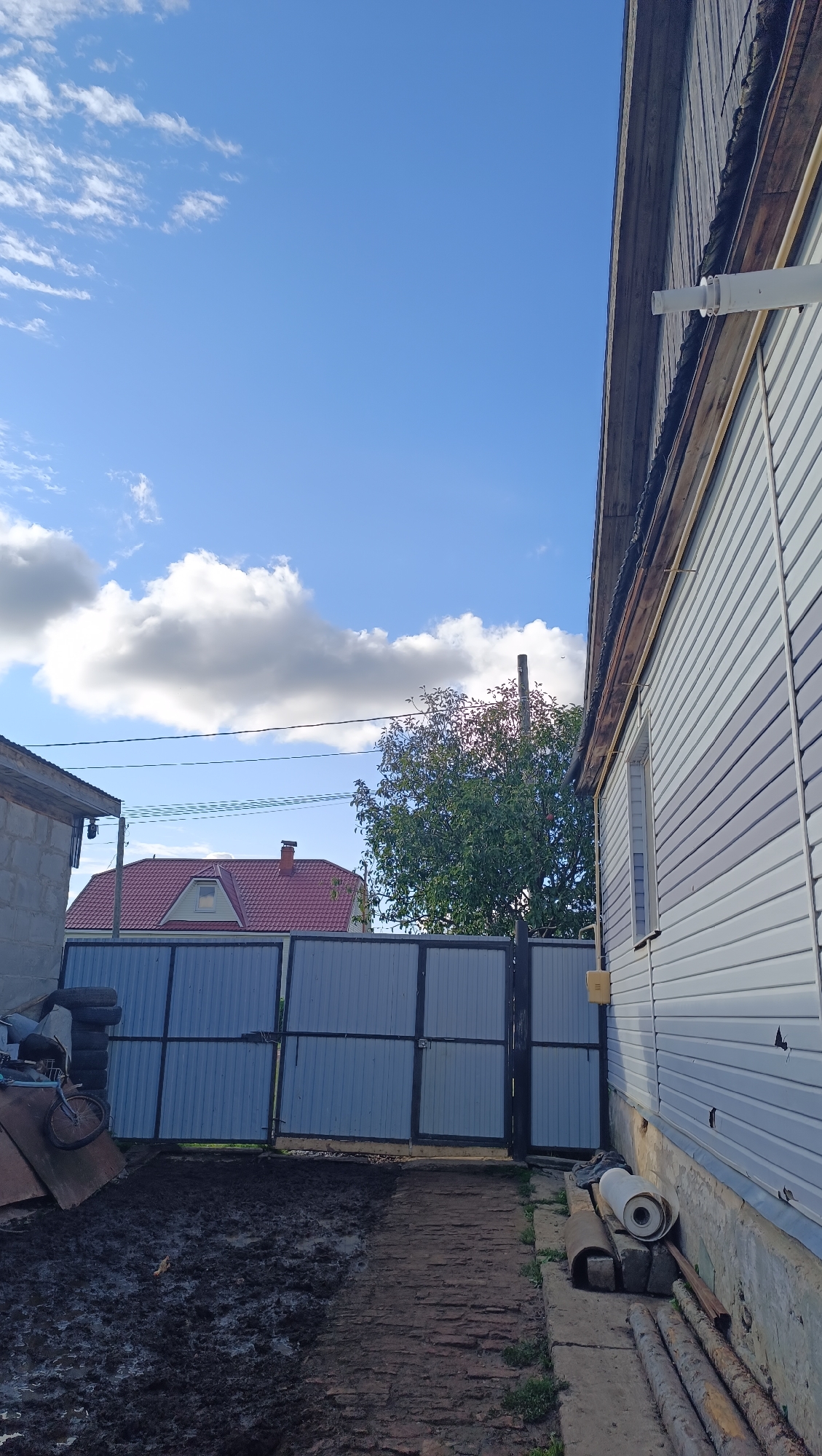 A vertical, wide shot captures a partially constructed or weathered rural scene under a bright blue sky with scattered white clouds. The foreground shows a muddy, uneven ground leading to a large, gray metal gate. To the left of the gate, stacked tires and a bicycle are visible near a low concrete structure. On the right side of the image, the exterior wall of a light-colored, horizontally paneled building dominates the frame, with a portion of a wooden-framed roof visible at the top. A white pipe extends from the building's wall outwards. In the background, behind the gate, a red-roofed house with a dormer window is visible, and a leafy green tree stands tall. A wooden utility pole is also present in the background. The scene appears to be a residential yard in a rural or semi-rural area, possibly Karsha, Russia, judging by the architecture and overall environment. There are no people visible in the image, and no discernible actions or interactions are taking place.