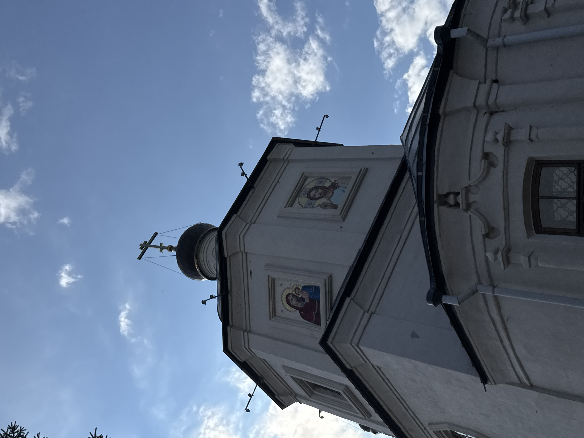 An exterior, low-angle, medium shot captures a portion of a white, octagonal church structure against a bright blue sky with scattered white clouds. The building features a dark dome topped with a gold cross. Two framed religious icons are visible on the facade: one depicting the Virgin Mary holding the infant Jesus, and another showing a male figure, likely Jesus Christ. The church has a dark, sloped roofline and decorative architectural elements. A tree branch is visible in the upper left corner. The scene is likely located in Kazan, Russia, as indicated by the architectural style and the presence of orthodox iconography.