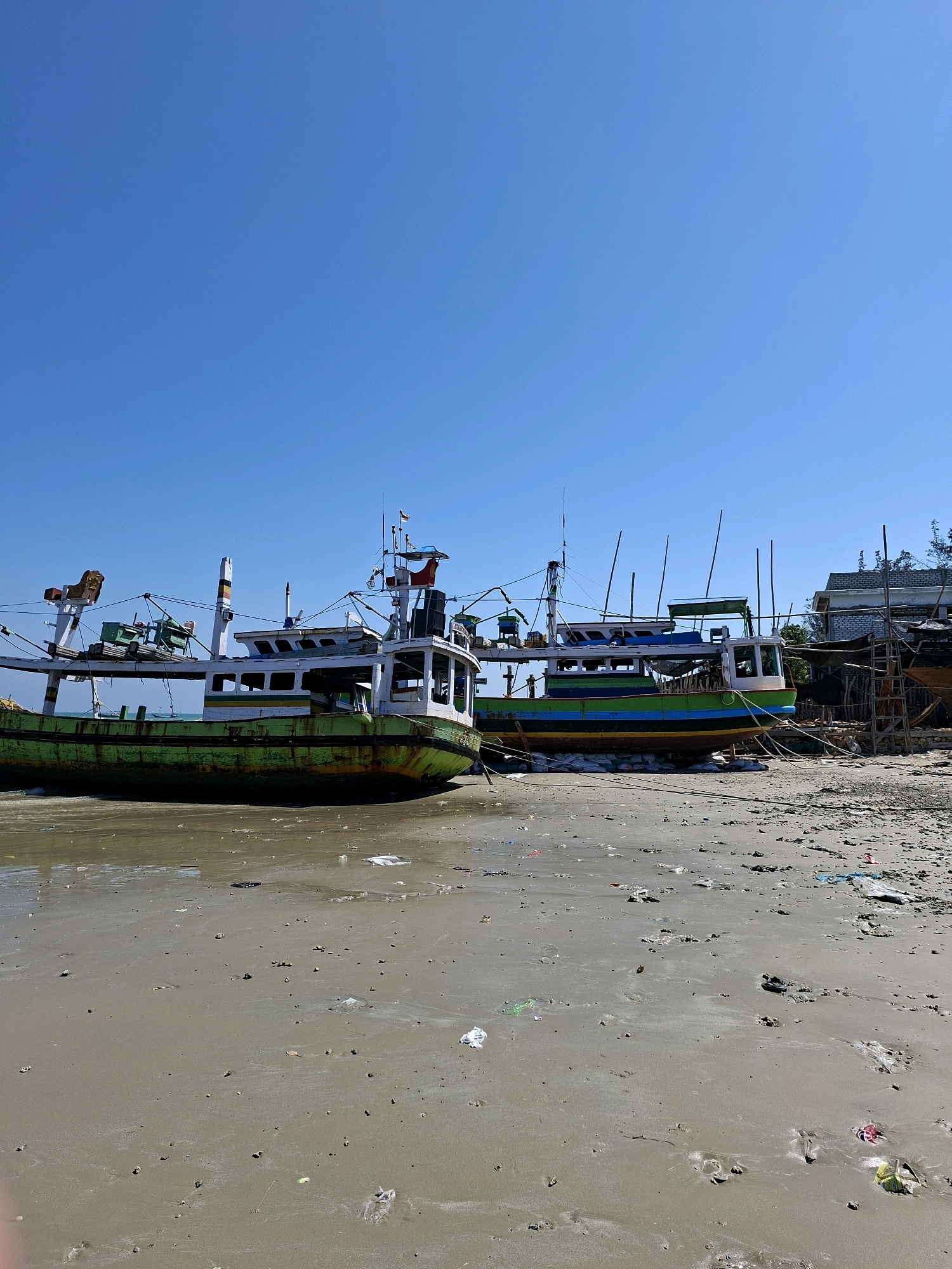 Two fishing boats are beached on a sandy shore under a clear blue sky. The boat in the foreground is primarily green with a white cabin and appears weathered. The second boat, slightly further back and to the right, has a blue and green hull with a white cabin. The shoreline is littered with various debris, including plastic bags and other trash. In the background, there are structures that appear to be part of a boatyard or maintenance area, with scaffolding and partially constructed or repaired boats. The scene is indicative of a coastal area in Camplong, Indonesia, likely a fishing village or port.