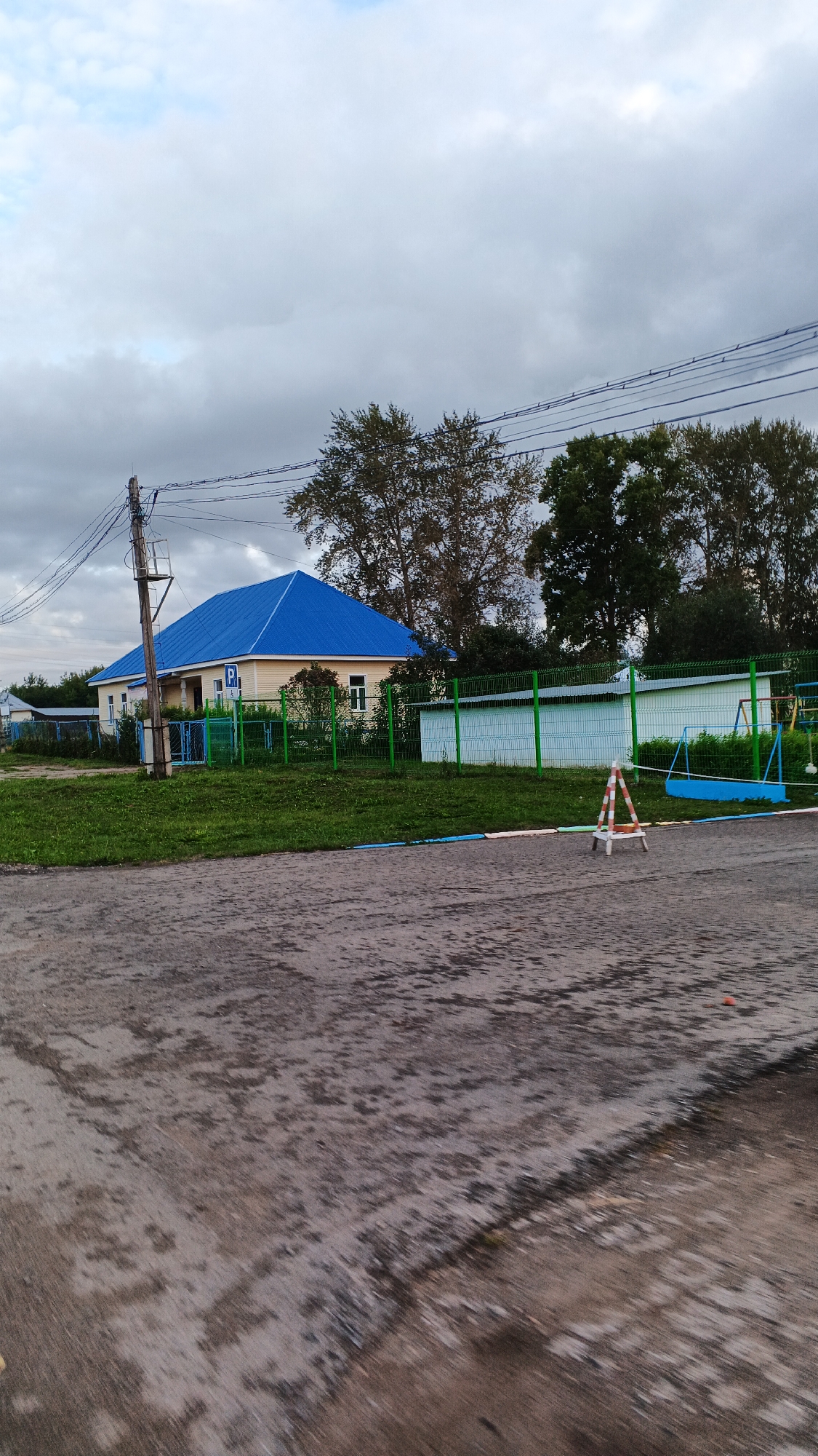 A light yellow, single-story building with a bright blue, gabled roof stands behind a green metal fence in a grassy area. The building has multiple windows and a sign with a blue "P" on it, indicating parking. To the right of the building, a white structure is visible behind the fence, and further to the right, a playground with a blue swing set is partially visible. A wooden utility pole with multiple wires is located to the left of the building. The foreground is a paved surface, possibly a road or parking lot, with visible water patches. A small, orange and white A-frame sign is placed on the paved surface in front of the fence. The sky is overcast with gray clouds. The scene appears to be located in the village of Podgornoye, Russia.