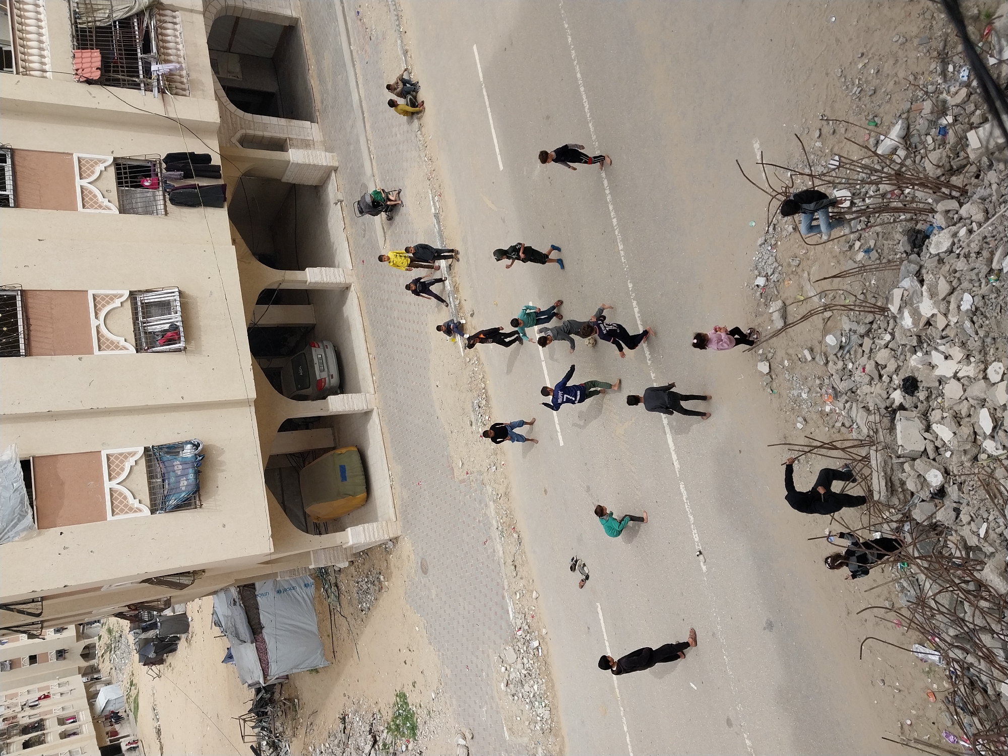 This high-angle image depicts a residential street scene in Khan Yunis, Palestinian Territory. In the immediate foreground, a significant mound of construction debris, consisting of concrete chunks and exposed, twisted rebar, borders the asphalt road. On this rubble, one individual in dark clothing crouches, while a young girl in a pink top and black pants stands nearby. Further right, another individual sits on a white object amidst the rebar.
On the gray asphalt road, approximately eight to ten barefoot children, predominantly boys, are actively playing football. One boy wears a blue jersey with "MBAPPE 7" on the back. Other children are visible along the road's edges; some sit on a sandy curb strewn with debris, while others stand observing the game. A dark-colored baby stroller is parked on the sidewalk. The road surface has faded white lane markings and its shoulders are composed of sand, dirt, and scattered refuse.
The mid-ground features a multi-story, light beige residential building on the right. This structure exhibits decorative arched window frames with grilles, and some windows are partially covered with plastic sheeting. Laundry, including dark garments and a pink item, hangs from upper-story windows. The ground level incorporates arched open spaces, providing shelter for a vehicle covered by a yellow tarp and a silver sedan. In the background and to the left, additional multi-story residential buildings are visible, alongside an open, sandy area containing multiple makeshift shelters constructed from tarps and corrugated metal, one displaying a "UNICEF" logo. The overall environment suggests a mix of residential activity amidst signs of urban deterioration or damage.