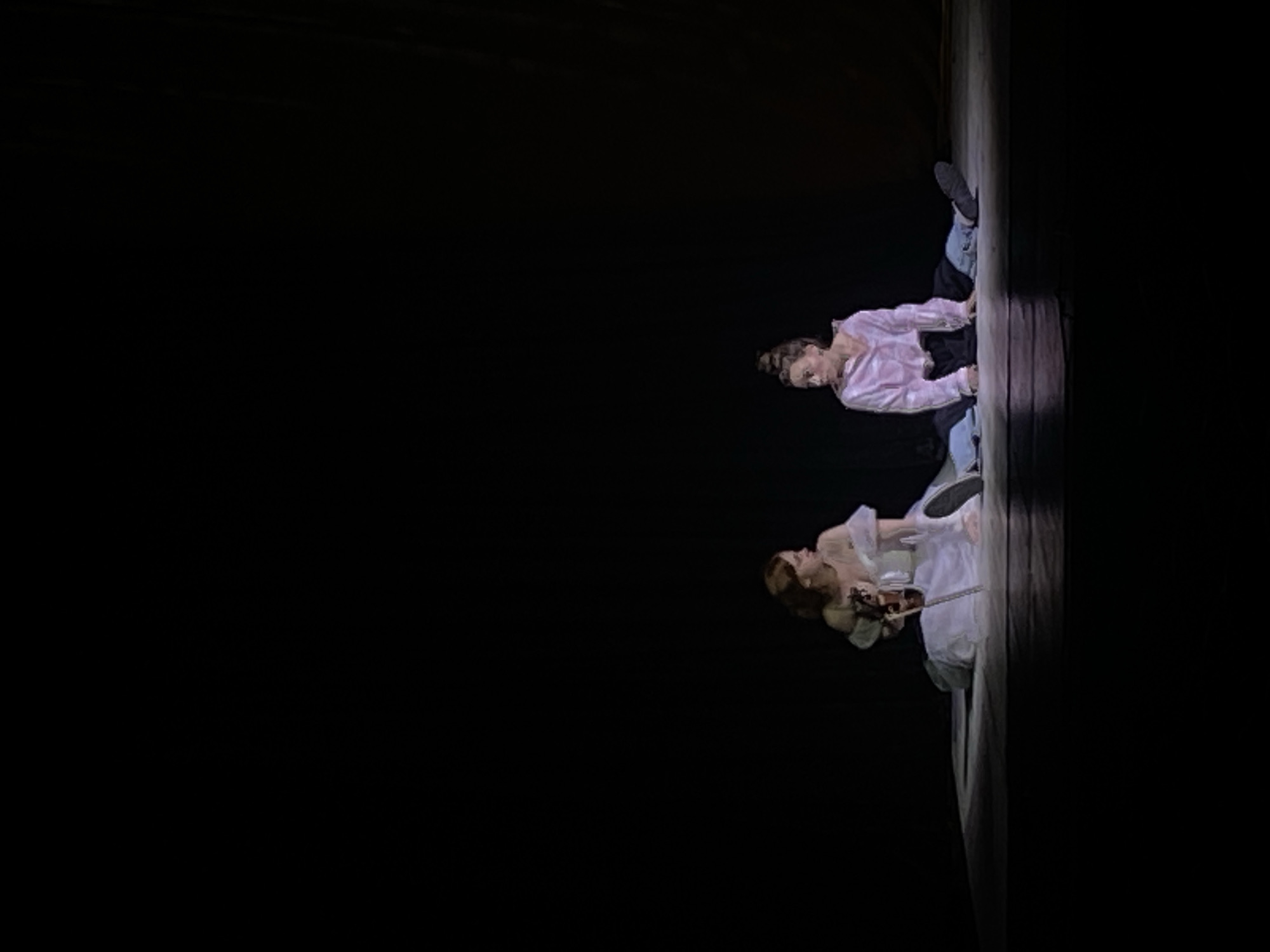 Two performers are on stage, one in a white gown playing a violin and the other in a pink shirt and jeans. They are in a dark theater setting with visible stage flooring. Location is unknown.