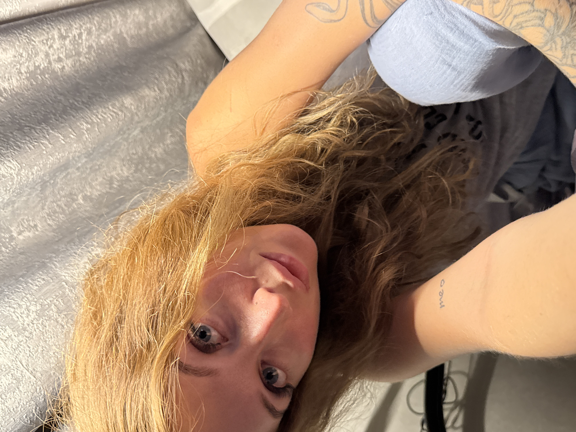 A close-up, eye-level shot focuses on a blonde woman with wavy hair. She is indoors, lying on a silver-gray, textured surface that appears to be bedding or furniture. Her head is slightly tilted. The woman's eyes are blue, and her skin is fair. Her left arm, visible in the shot, has a tattoo of the word "meo" in small cursive letters on the inner wrist. The arm also has a tattoo. She is wearing a light gray shirt. The location in the city of Chaikovsky, Russia cannot be confirmed.