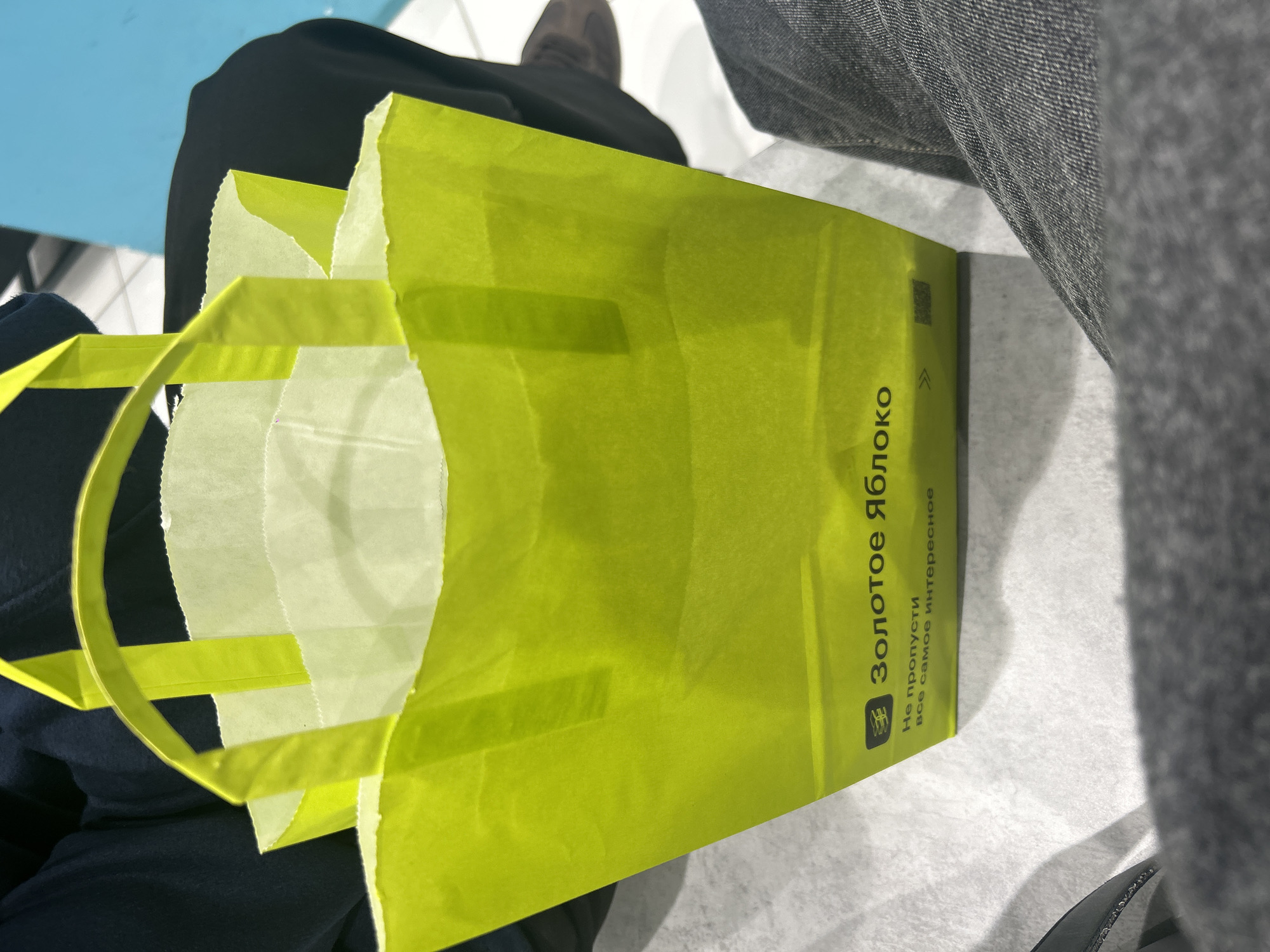 The media file captures an indoor scene in what appears to be a public space, such as a shopping mall or waiting area in Saint Petersburg, Russia. The primary subject is a vibrant, open, lime-green shopping bag, likely made of paper or thin cardboard, featuring matching handles. Inside the bag, layers of translucent white tissue paper are visible.
The shopping bag prominently displays text in Cyrillic script. The main branding reads "Золотое яблоко" (Zolotoe yabloko), which translates to "Golden Apple," identifying it as from a well-known Russian beauty and cosmetics chain. Below this, smaller text says "Не пропусти" (Don't miss out) and "все самое интересное" (all the most interesting things). A small square symbol, possibly a QR code, and a right-pointing arrow are also visible on the bag.
The bag rests on or near a person's lap. Parts of dark clothing (possibly pants or a skirt) are visible on the left, and gray fabric, likely jeans or trousers, is seen on the right. In the upper background, a dark shoe is partially visible, along with glimpses of a light blue surface (possibly seating or a table) and light-colored flooring. The photograph is taken from a slightly elevated perspective, looking down into the bag. The lighting is bright and even, indicating a daytime setting. The image depicts a casual moment, likely after a shopping experience.