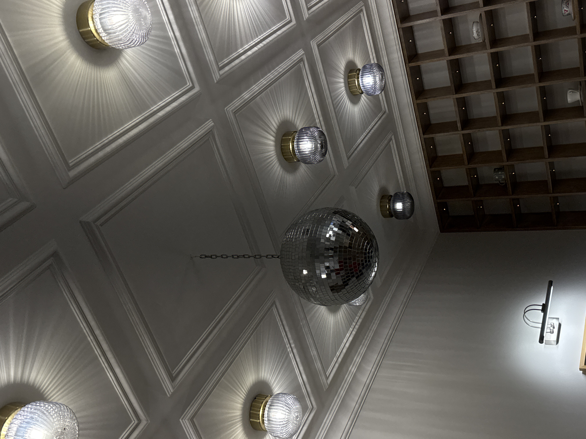A photograph taken indoors in Yerevan, Armenia, reveals a complex interplay of architectural and decorative elements. The lower left corner shows a light fixture with a ribbed glass cover and a gold base, mounted on a white paneled wall. The wall is structured with geometric, recessed paneling. Moving upwards and towards the center, a disco ball hangs from a chain, positioned amidst the paneled ceiling. Additional light fixtures of similar design punctuate the panels, creating a rhythmic pattern. To the right, a modern wall sconce is visible against a white wall. A wooden shelving unit with square compartments is overhead and to the upper right of the frame.