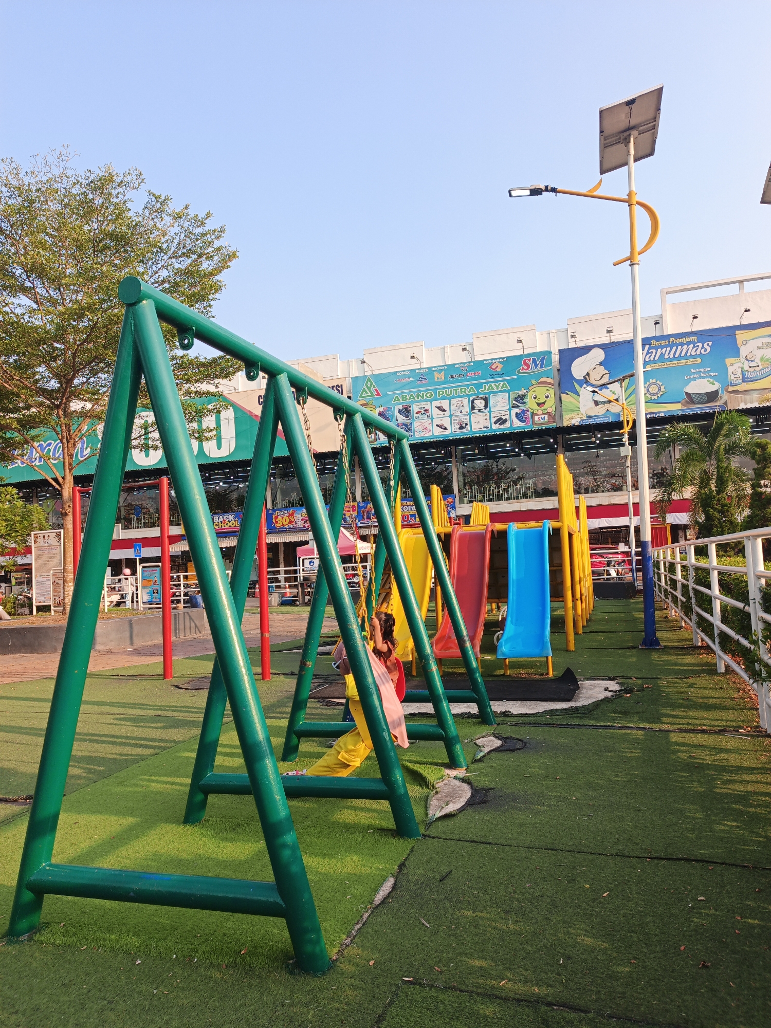 This is a full-shot, outdoor, daytime photograph of a playground with a swing set in the foreground. The swing set is painted green and has two yellow swings. One swing is occupied by a young girl wearing a pink shirt and yellow pants. The playground features artificial grass and several colorful slides, including red, yellow, and blue ones, arranged in a row. In the background, there is a building with large advertisements on its facade. One advertisement prominently features the text "Harumas" with an image of a chef. Other advertisements display various products and brands, along with the "SM" logo. A solar-powered street lamp is visible on the right side of the image, mounted on a pole with a yellow accent. A white fence runs along the right edge of the playground. The sky is clear and blue, indicating good weather. The presence of the "SM" logo and the architectural style of the buildings suggest this playground is likely located in a commercial area or shopping mall complex in Batam, Indonesia, possibly within the Nagoya Hill Shopping Mall area, which is known for its retail and entertainment facilities.